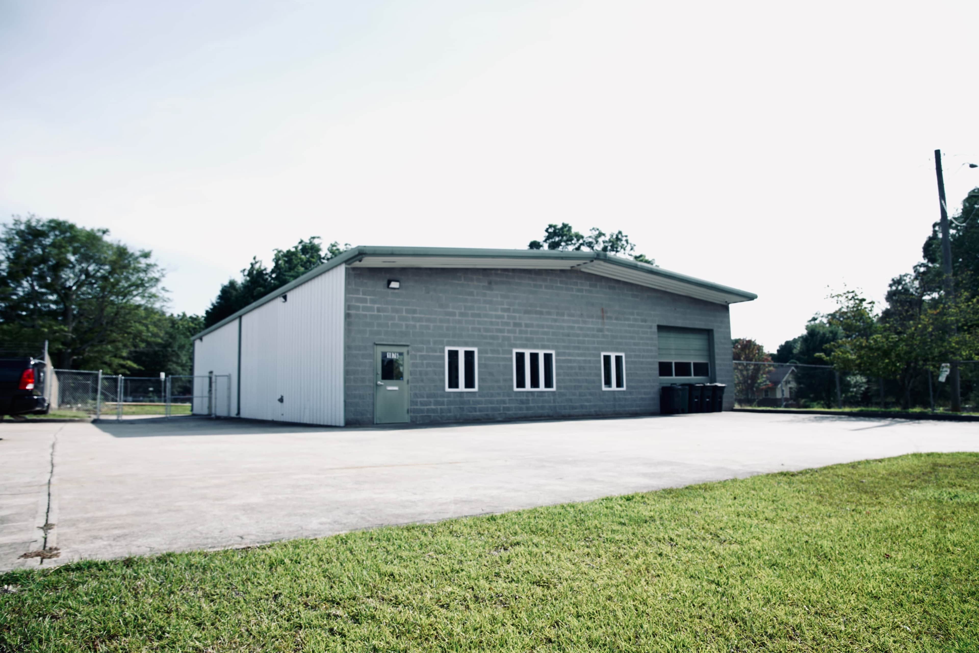 The image shows a single-story gray industrial building with a concrete parking area and some trees in the background.