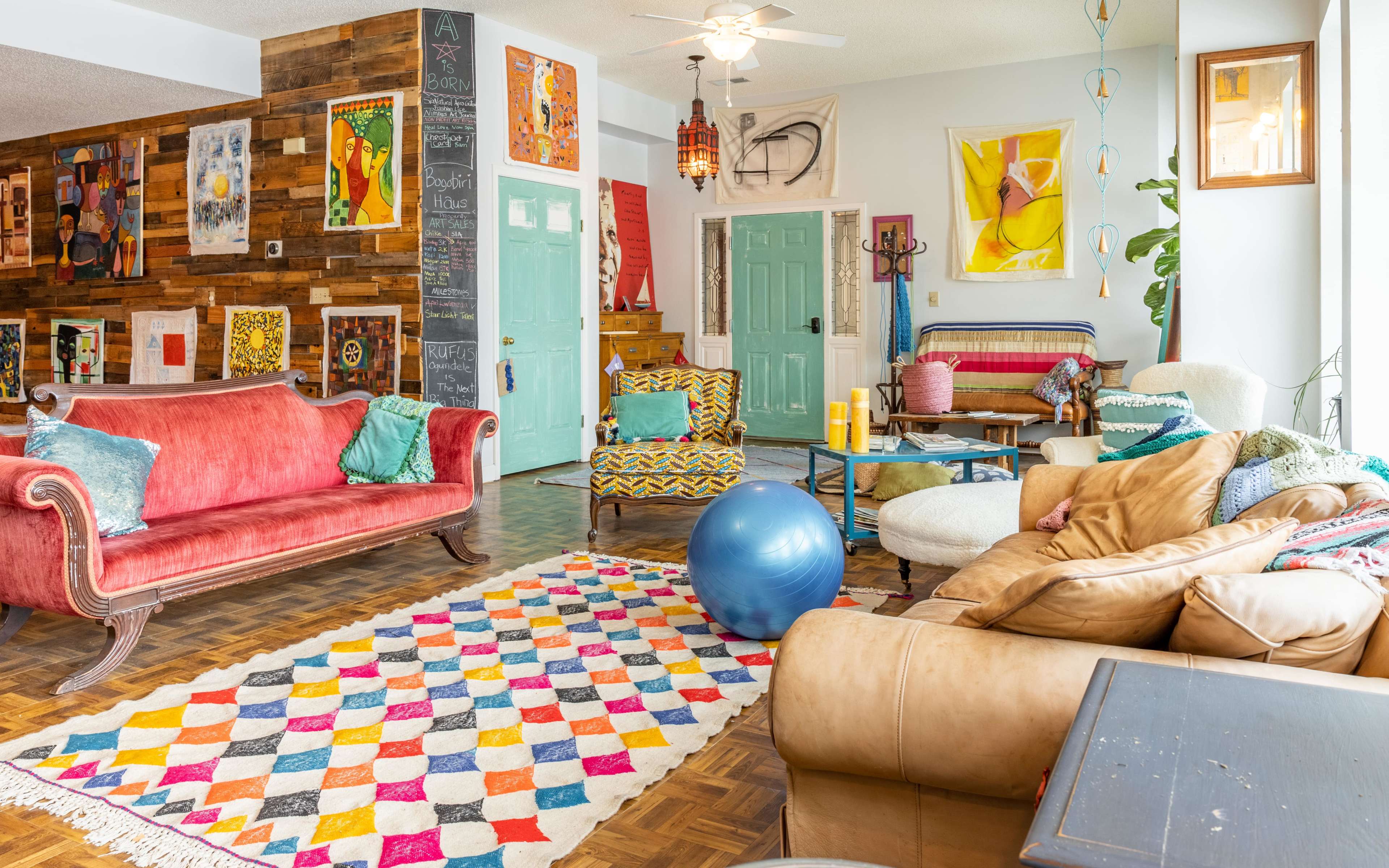 The scene features a colorful living room with various art pieces displayed on the walls, a patterned rug on the floor, and a mix of furniture including sofas and a stability ball.