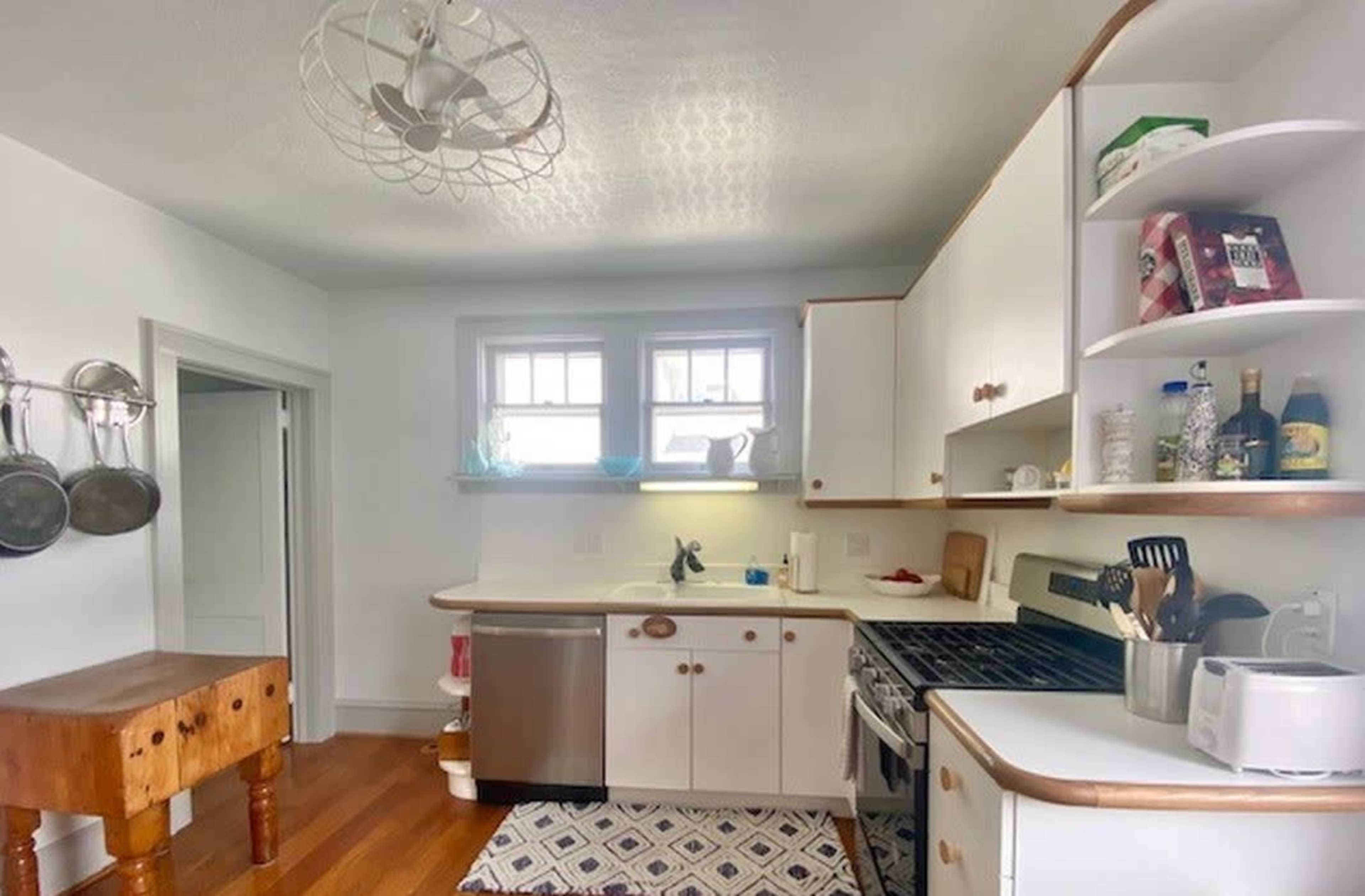 A tidy kitchen features a wooden prep table, stainless steel appliances, and white cabinetry lined with dishes and cooking supplies.