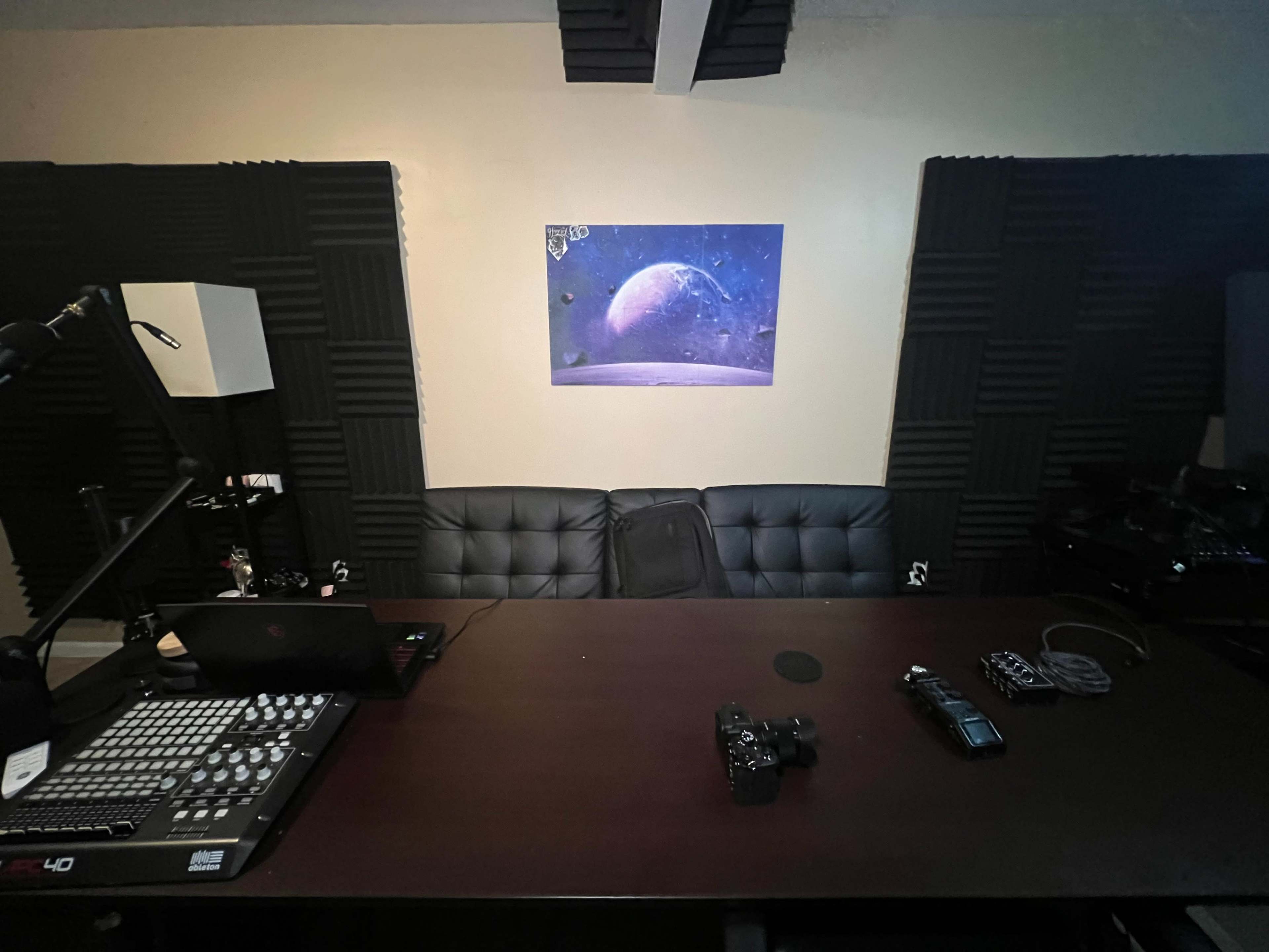A dimly lit recording studio setup with a desk, audio equipment, and a large print of a planet hanging on the wall.