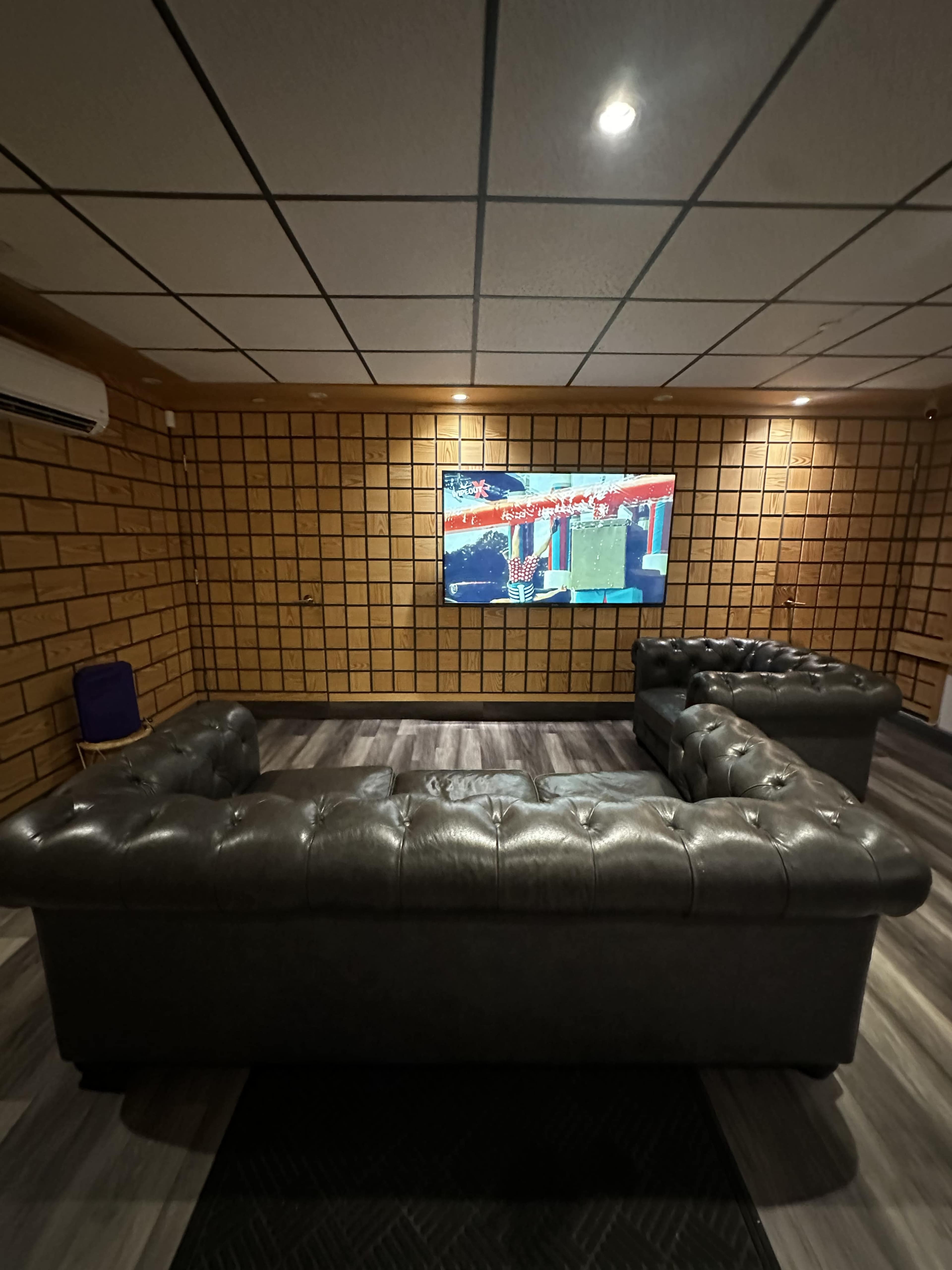 The room features a modern entertainment area with a sectional leather couch facing a large wall-mounted television and textured wooden paneling on the walls.