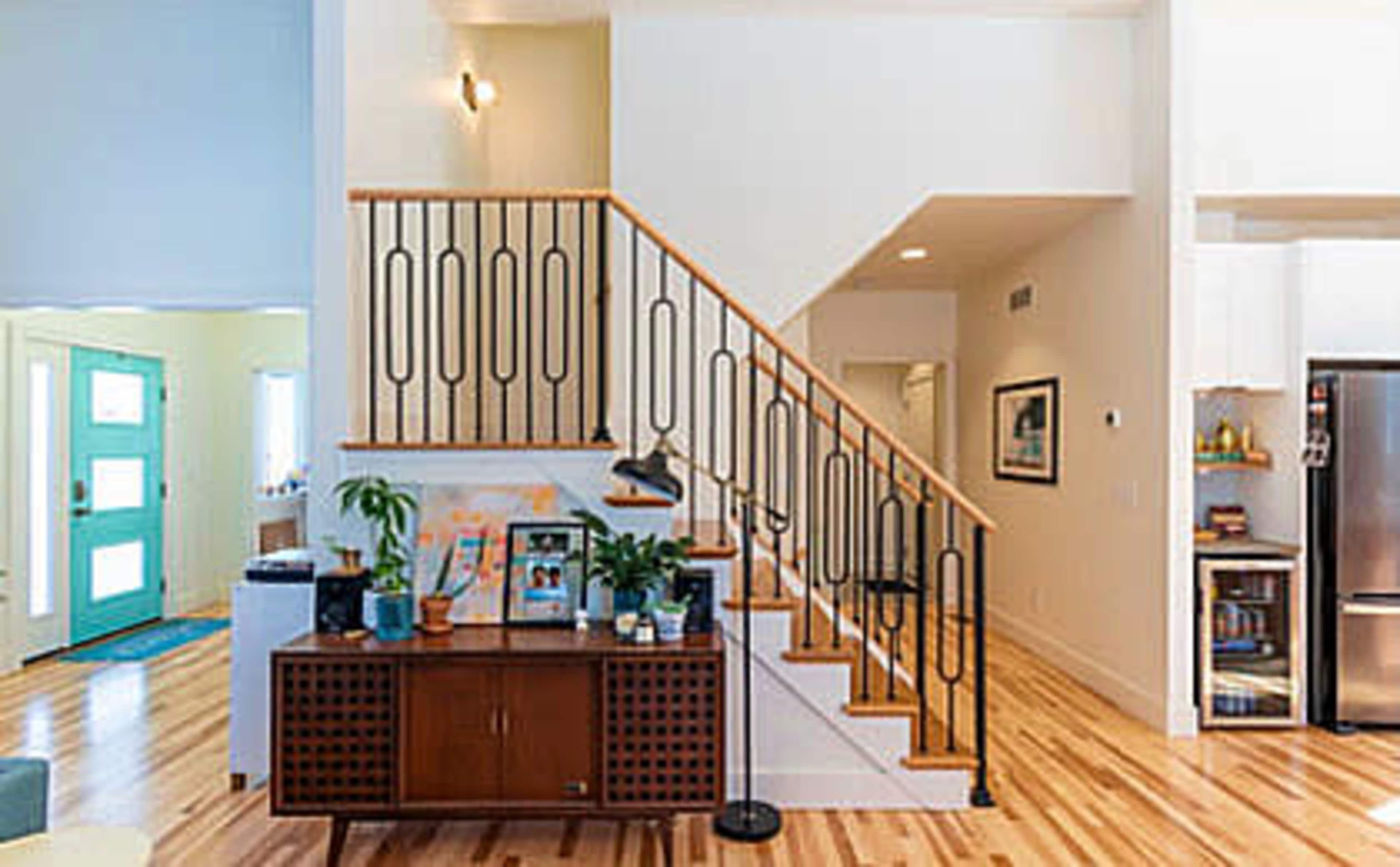 A modern interior featuring a staircase with decorative railing, a wooden cabinet with plants and decor, and an entrance with a turquoise door.