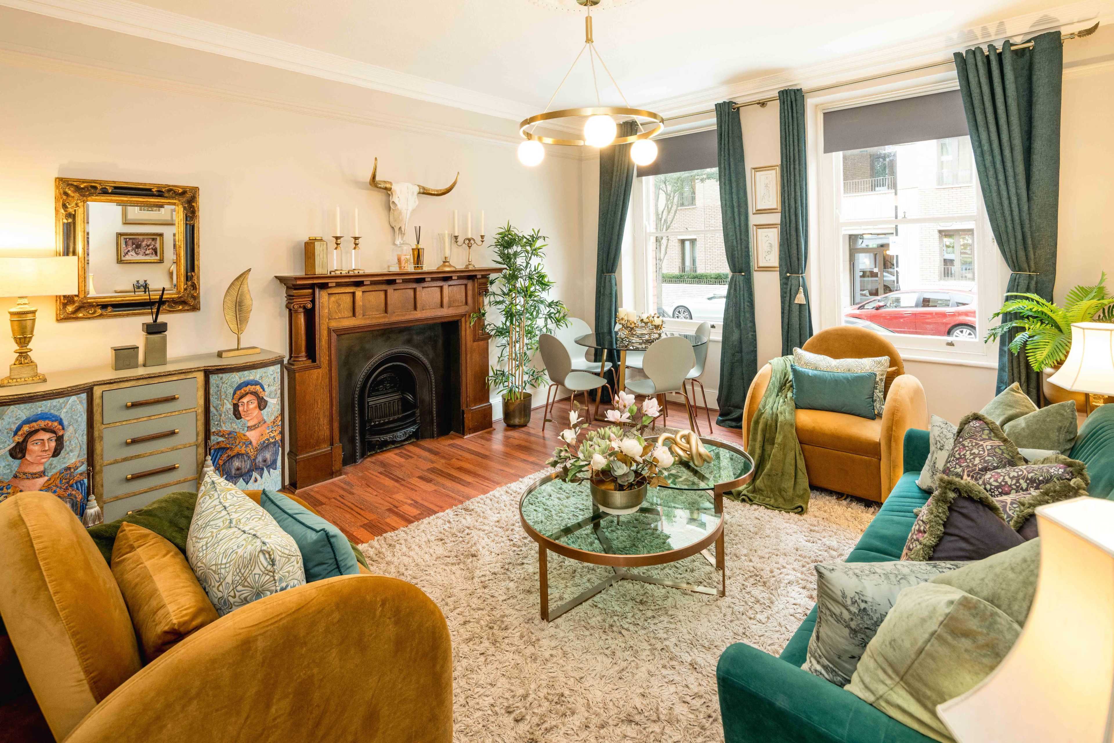 The image shows a stylish living room featuring a fireplace, a round glass coffee table, two comfortable sofas, and a dining area with chairs near a window.
