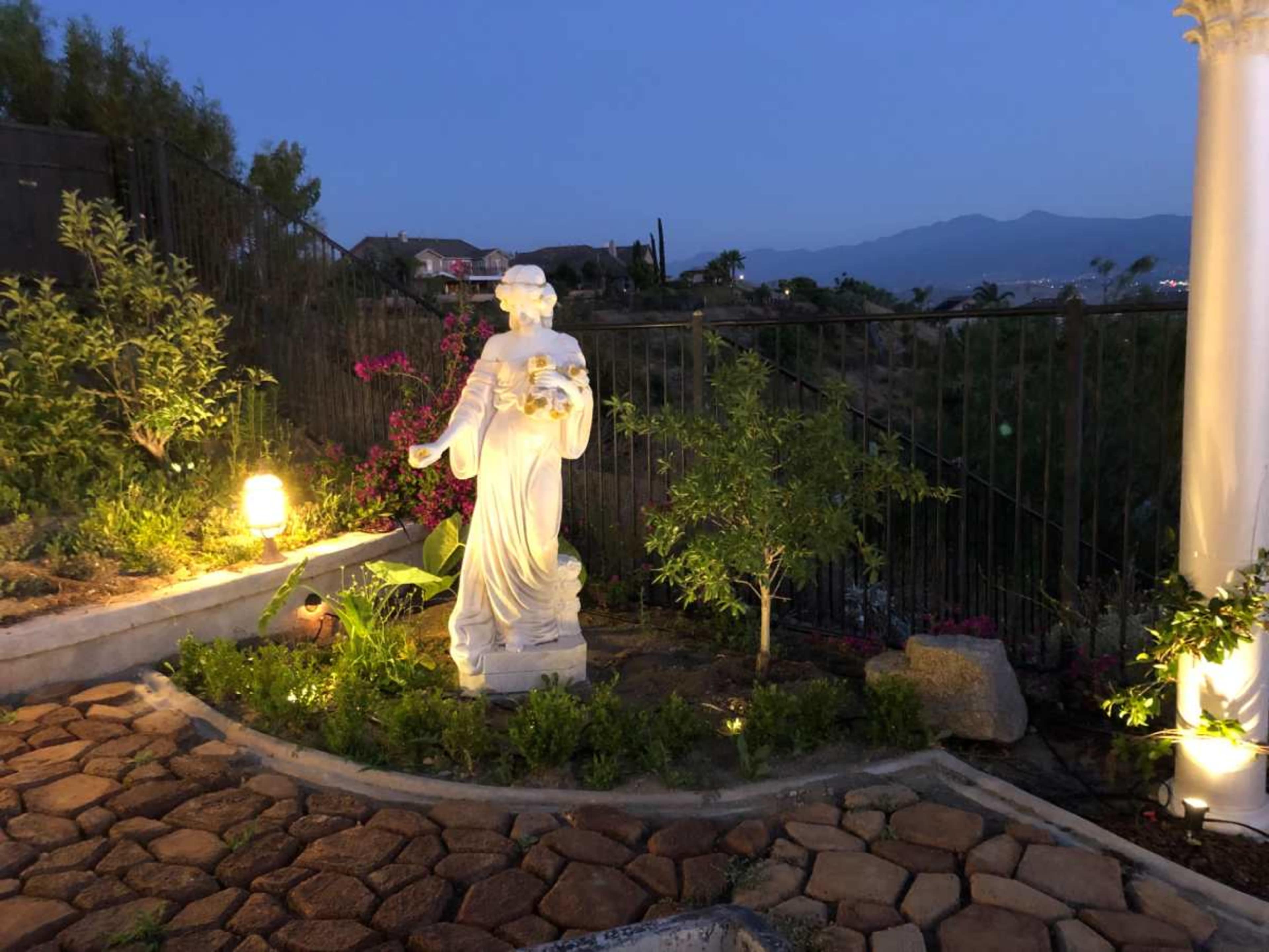 A white statue of a woman holding flowers stands in a garden illuminated by soft lights, surrounded by greenery and stone pathways.
