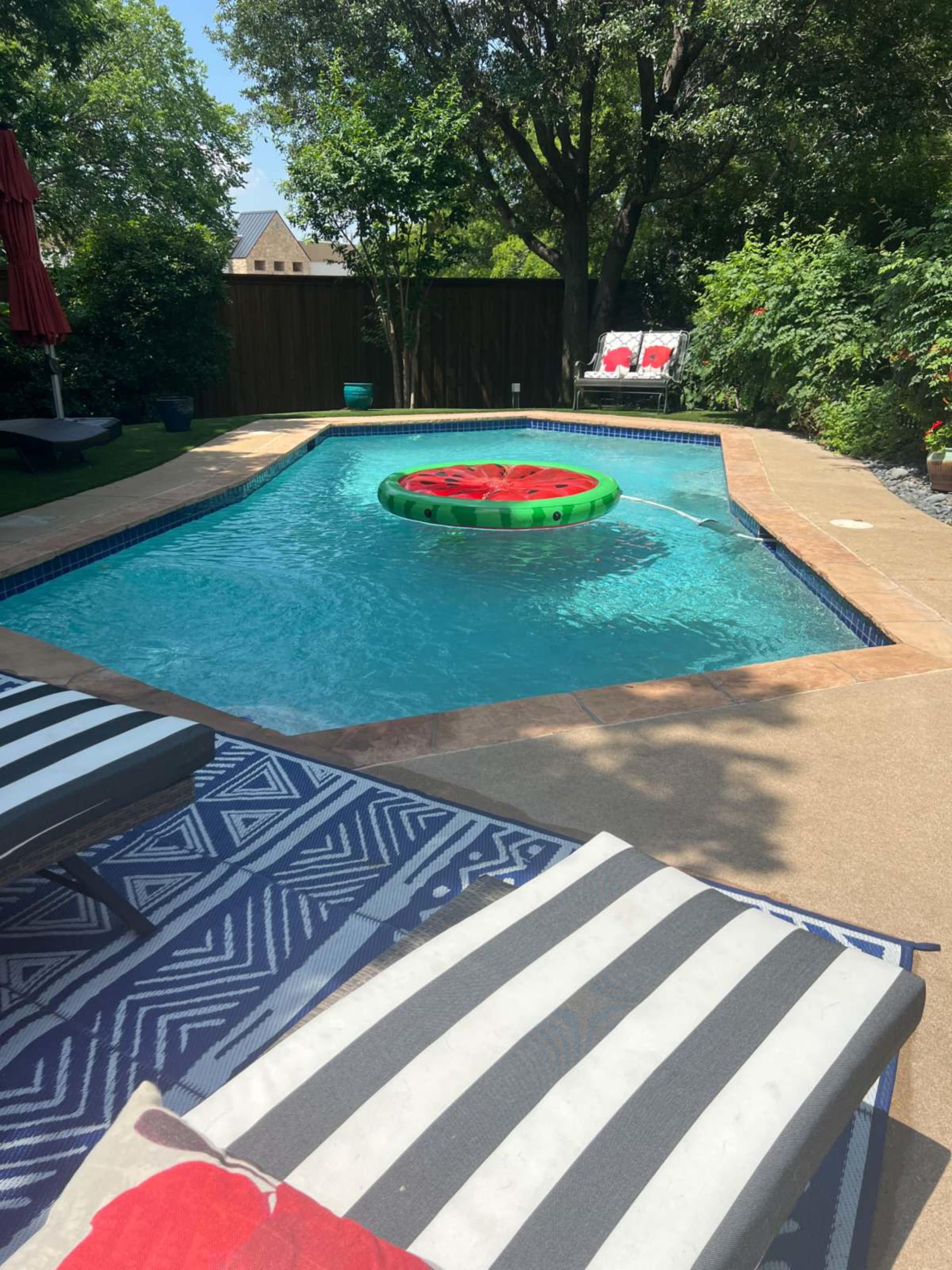 A swimming pool features a large green and red inflatable watermelon floating in the water, surrounded by striped lounge chairs and lush greenery.