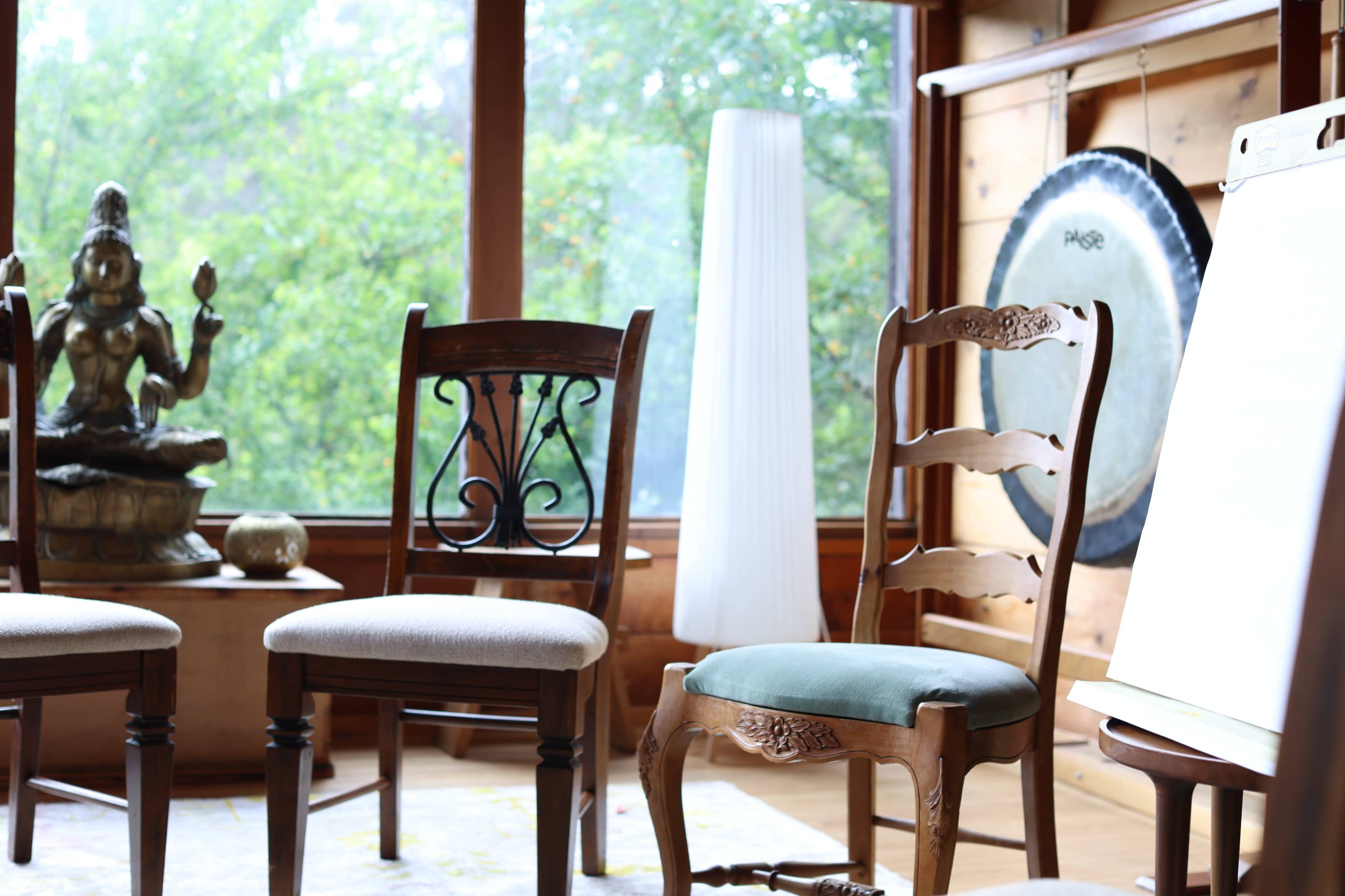 The image shows a cozy interior space with wooden chairs, a statue, a lamp, and a gong against a backdrop of large windows.
