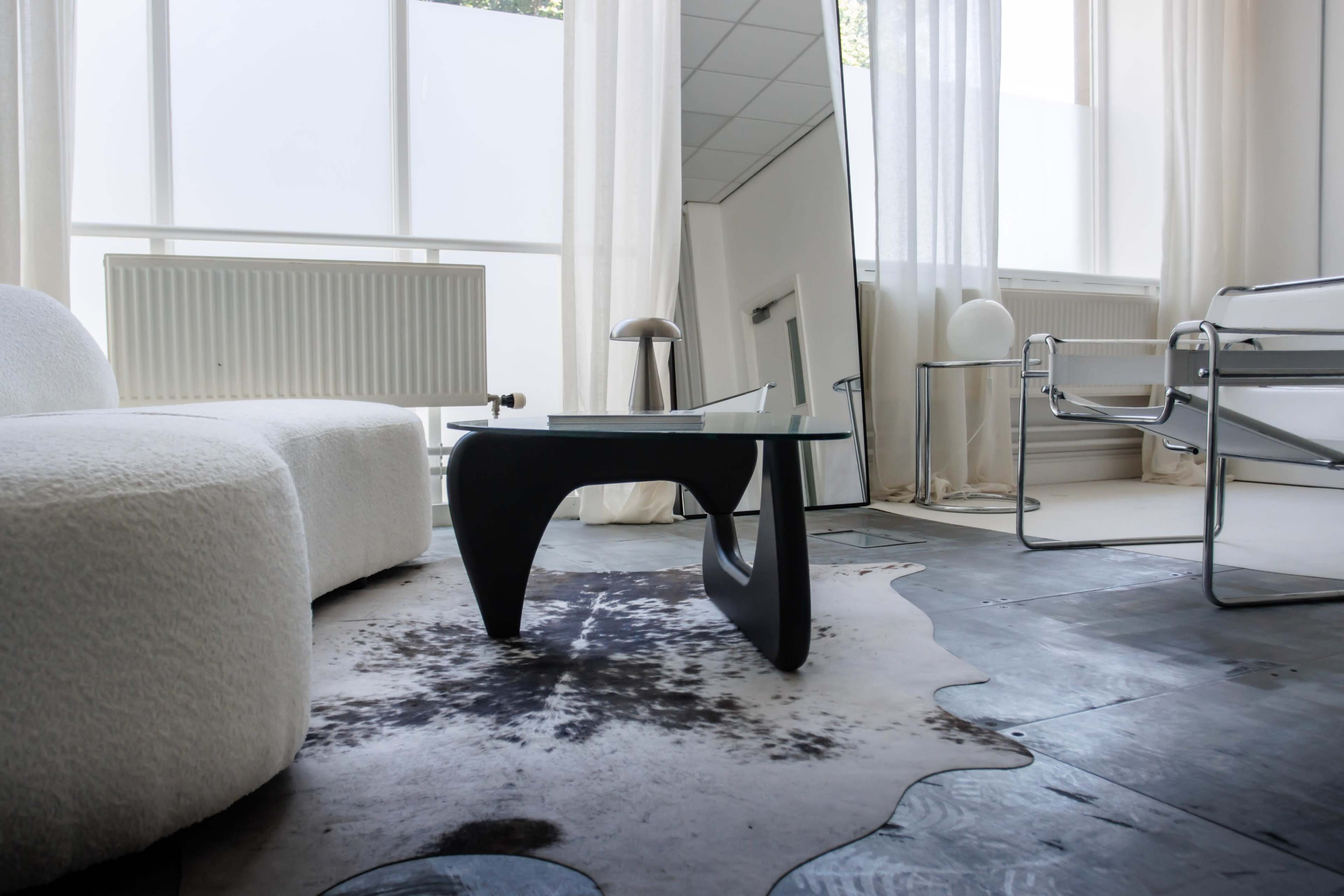 A modern living room featuring a white sofa, a glass coffee table with a black base, and a large mirror reflecting the space, complemented by natural light from large windows.