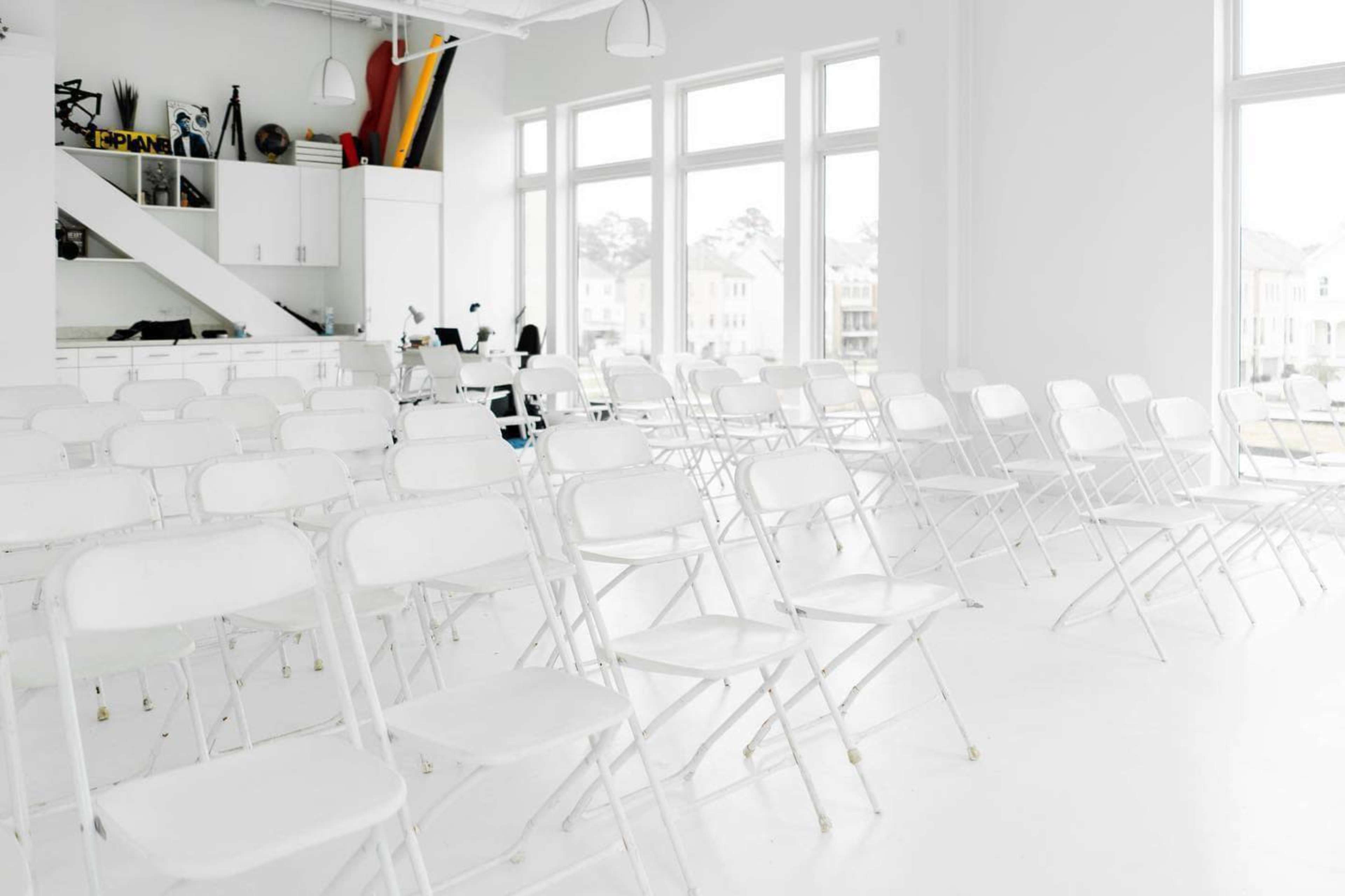 A bright, minimalist room filled with rows of white folding chairs and large windows allowing natural light to enter.