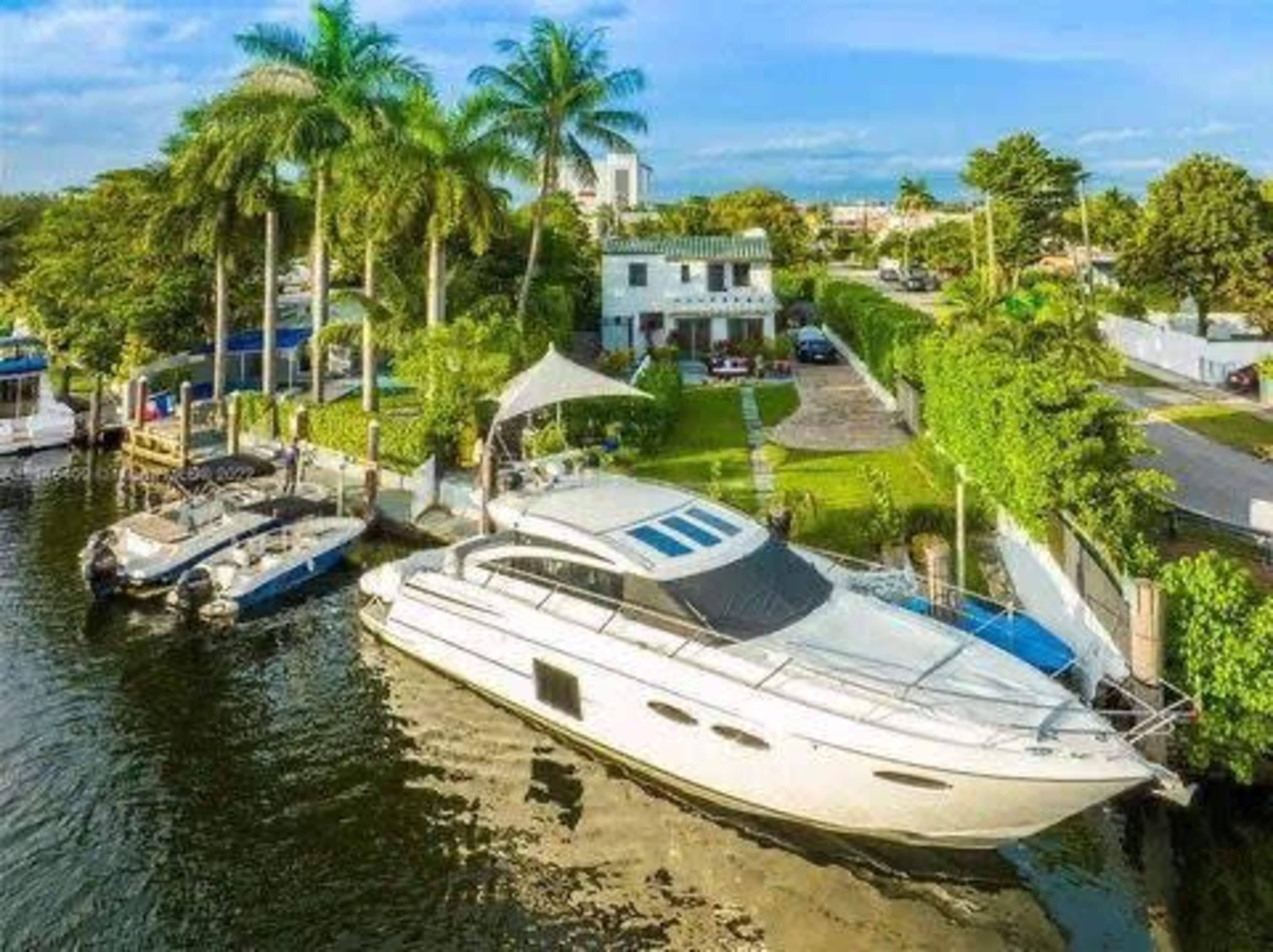 The image depicts a large white yacht docked beside a lush waterfront property, surrounded by palm trees and a well-maintained garden.
