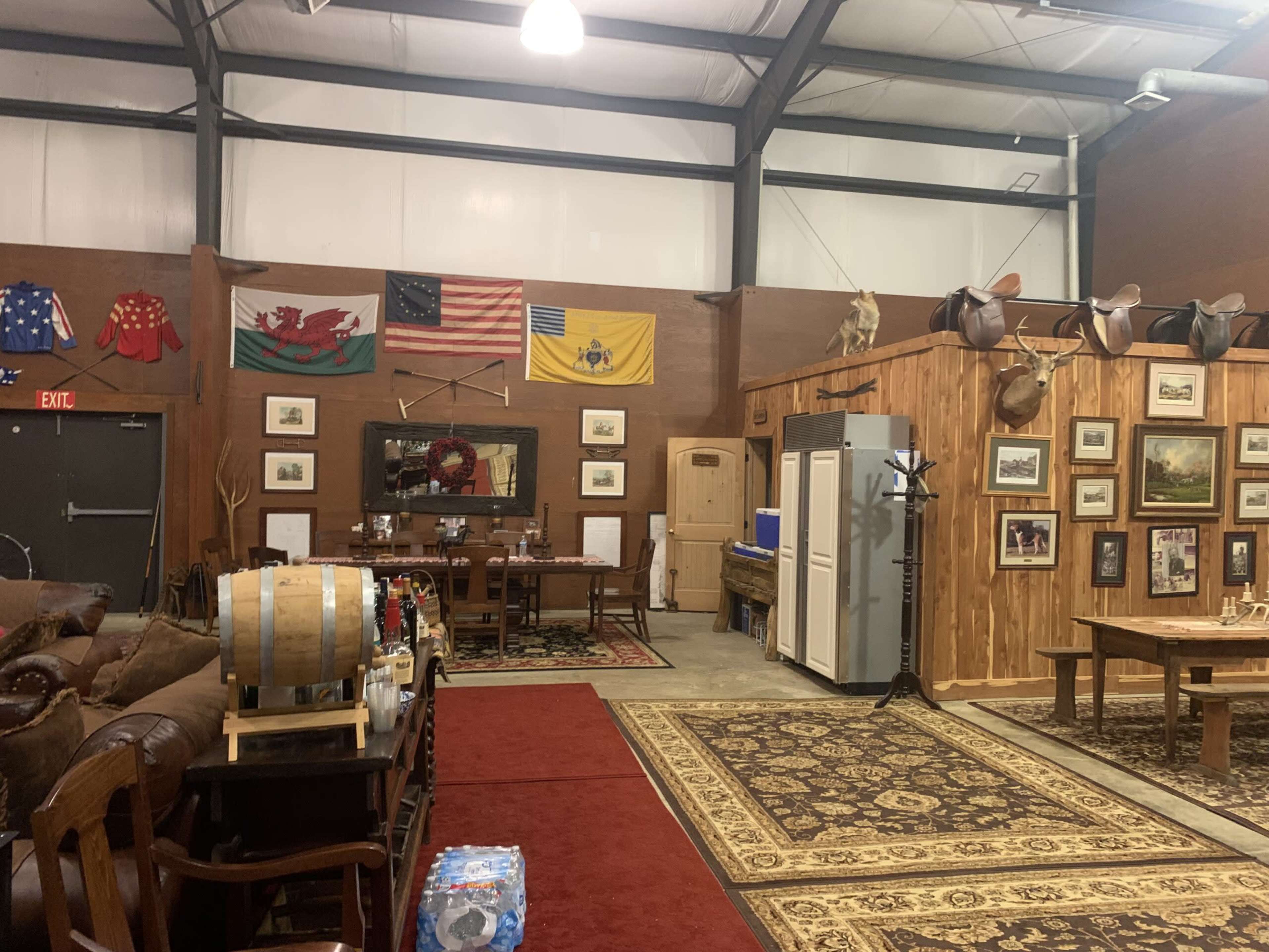 The scene depicts a spacious room with wooden walls, a dining area featuring a long table and chairs, several framed artworks, and decorative flags hanging above, along with seating arrangements and a faux rustic barrel.