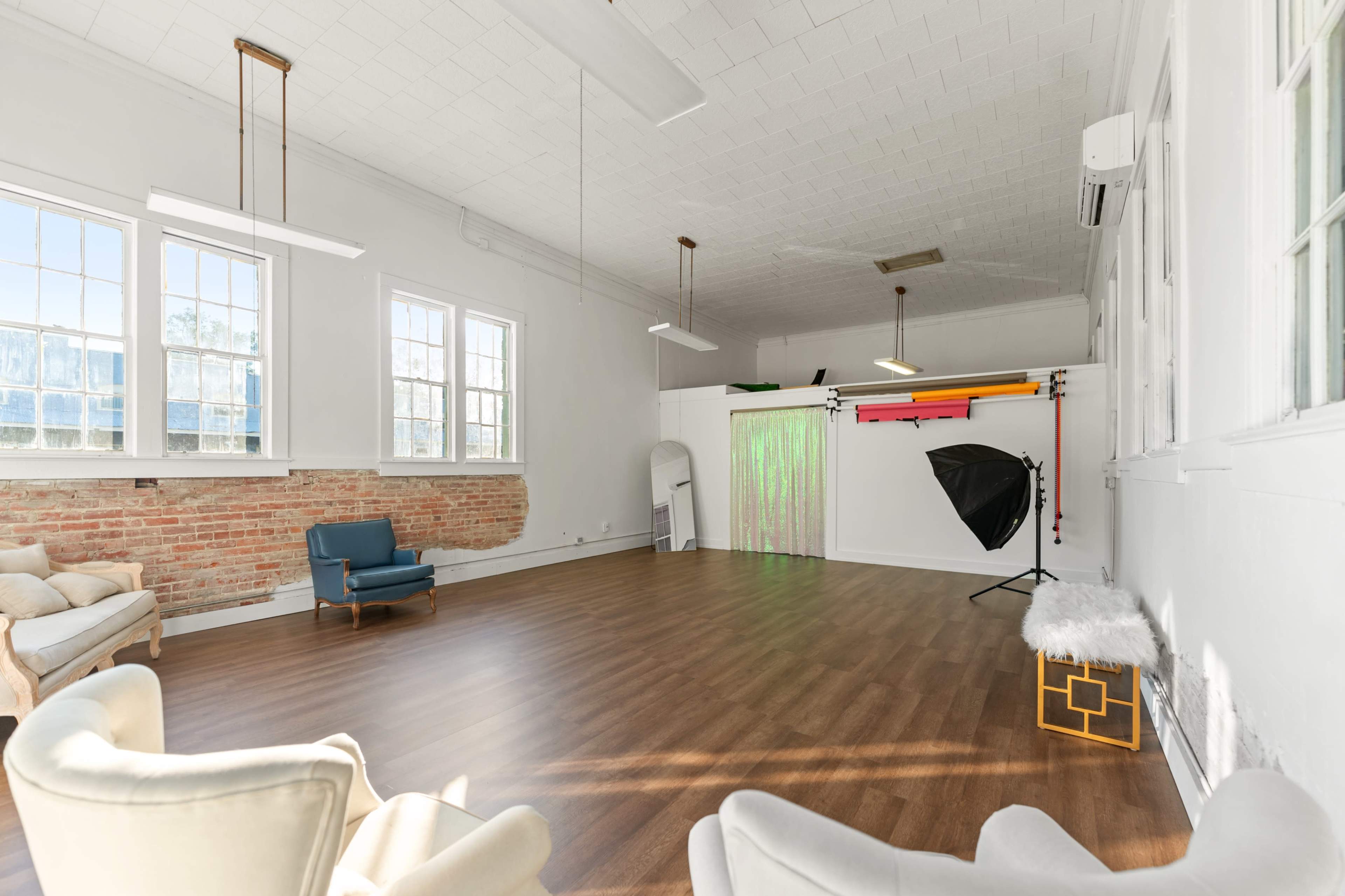 The image shows an empty, well-lit studio space featuring a wooden floor, large windows, and a backdrop setup for photography.