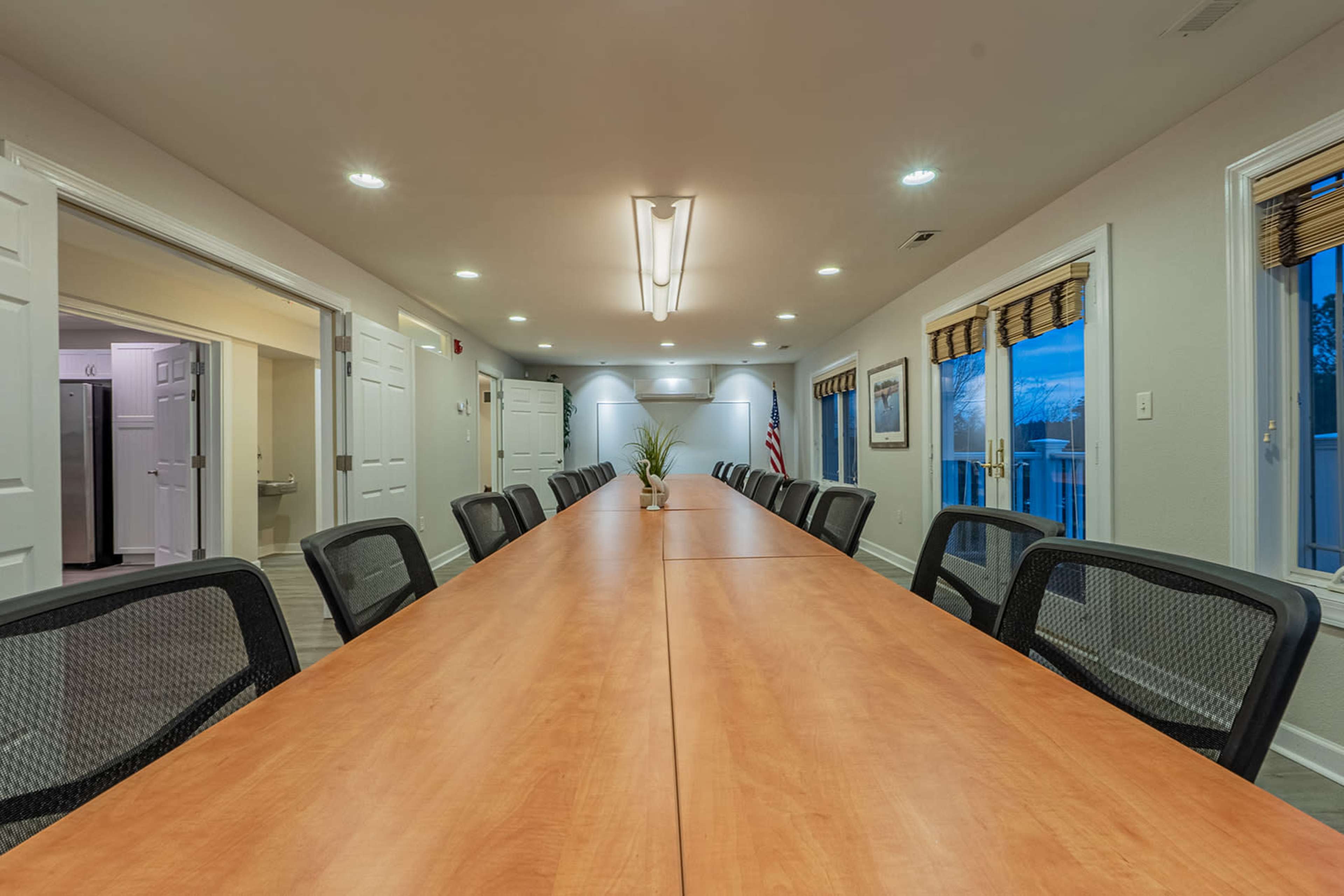 A long wooden conference table is set up in a well-lit meeting room with chairs arranged along each side and windows providing natural light.