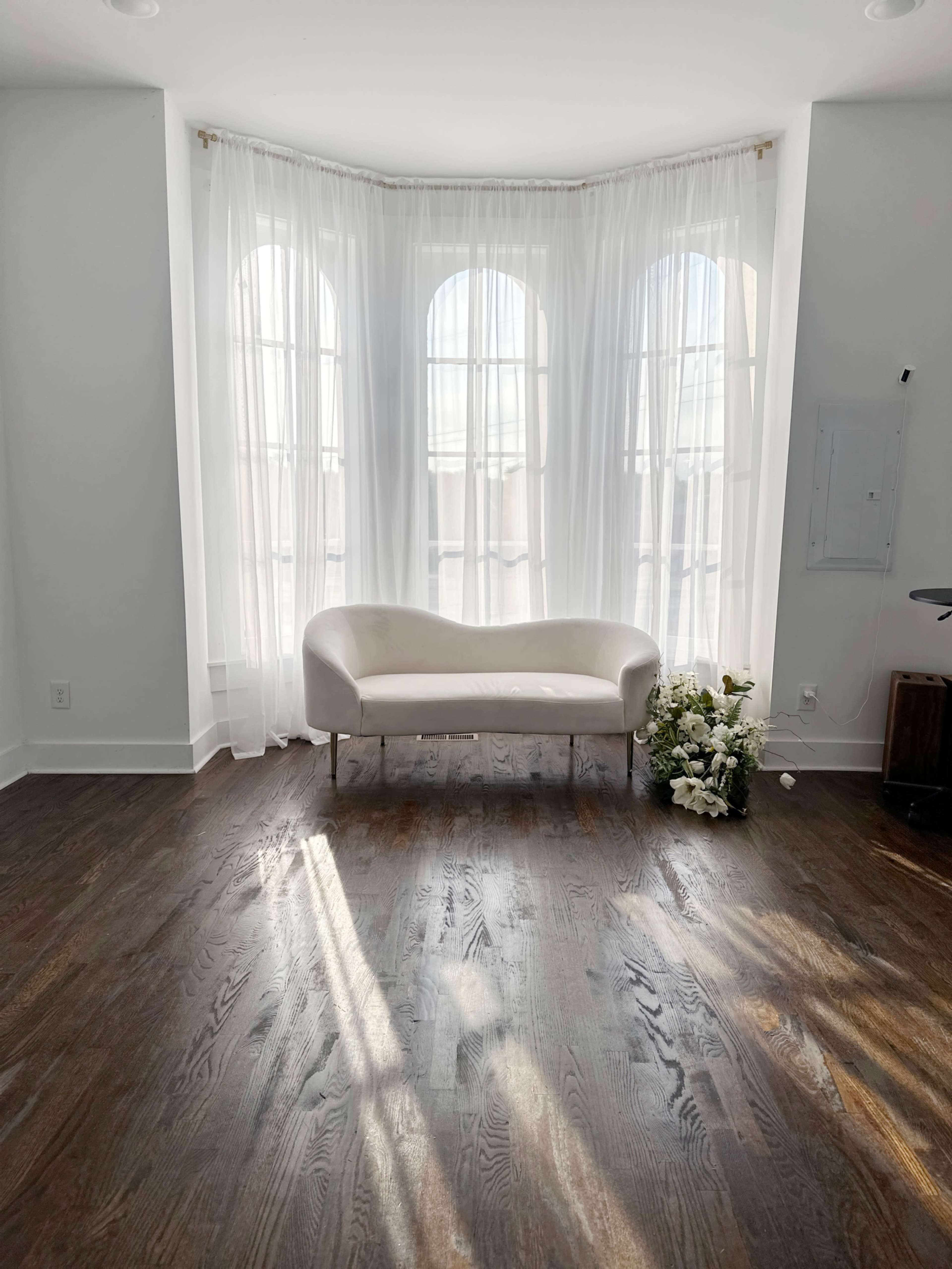 A white, curved sofa is positioned in a bright room with large windows dressed in sheer white curtains, and a bouquet of flowers rests on the wooden floor nearby.