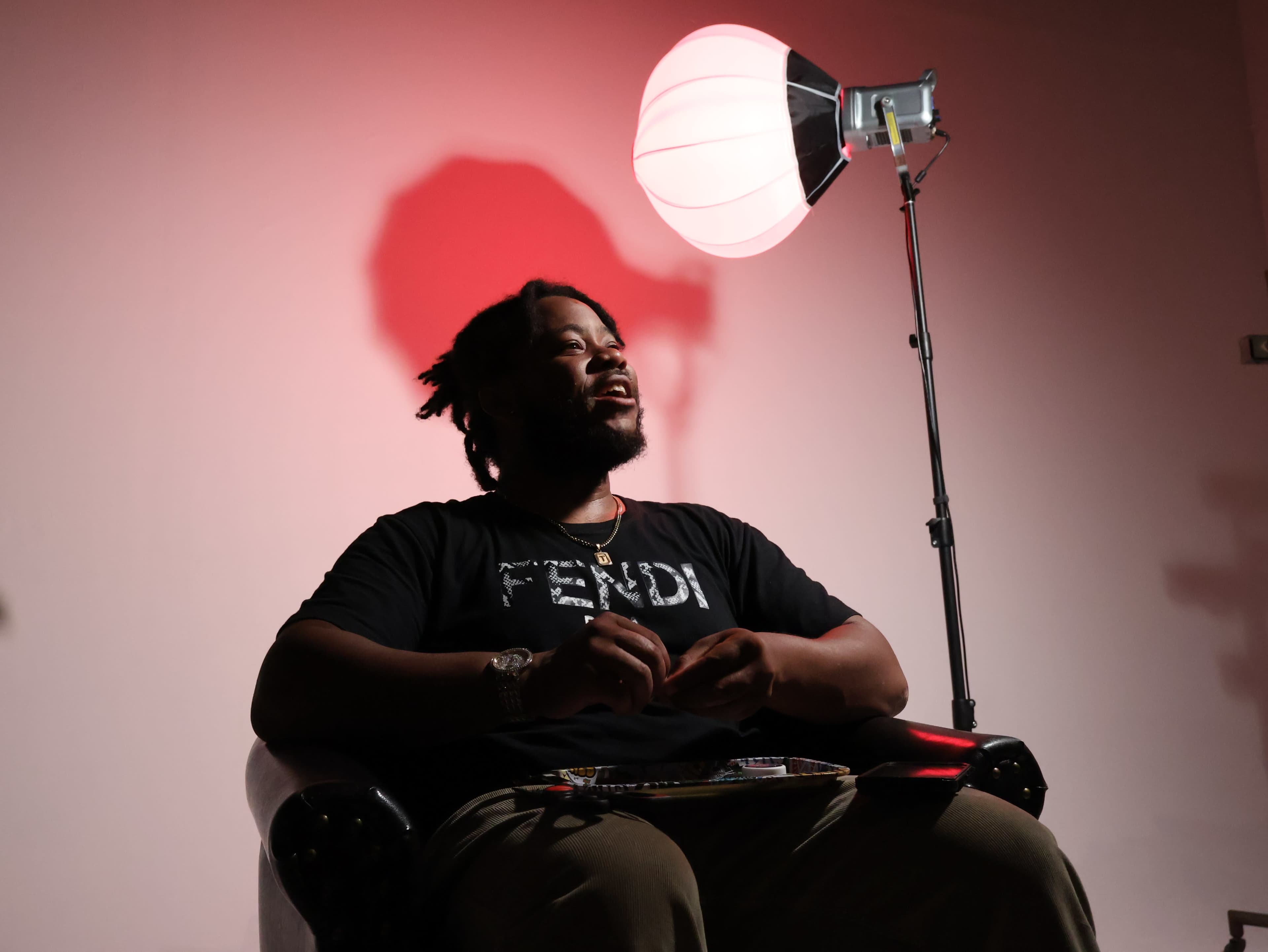 A man sits in a chair under a lamp that casts a red shadow, appearing engaged in conversation.