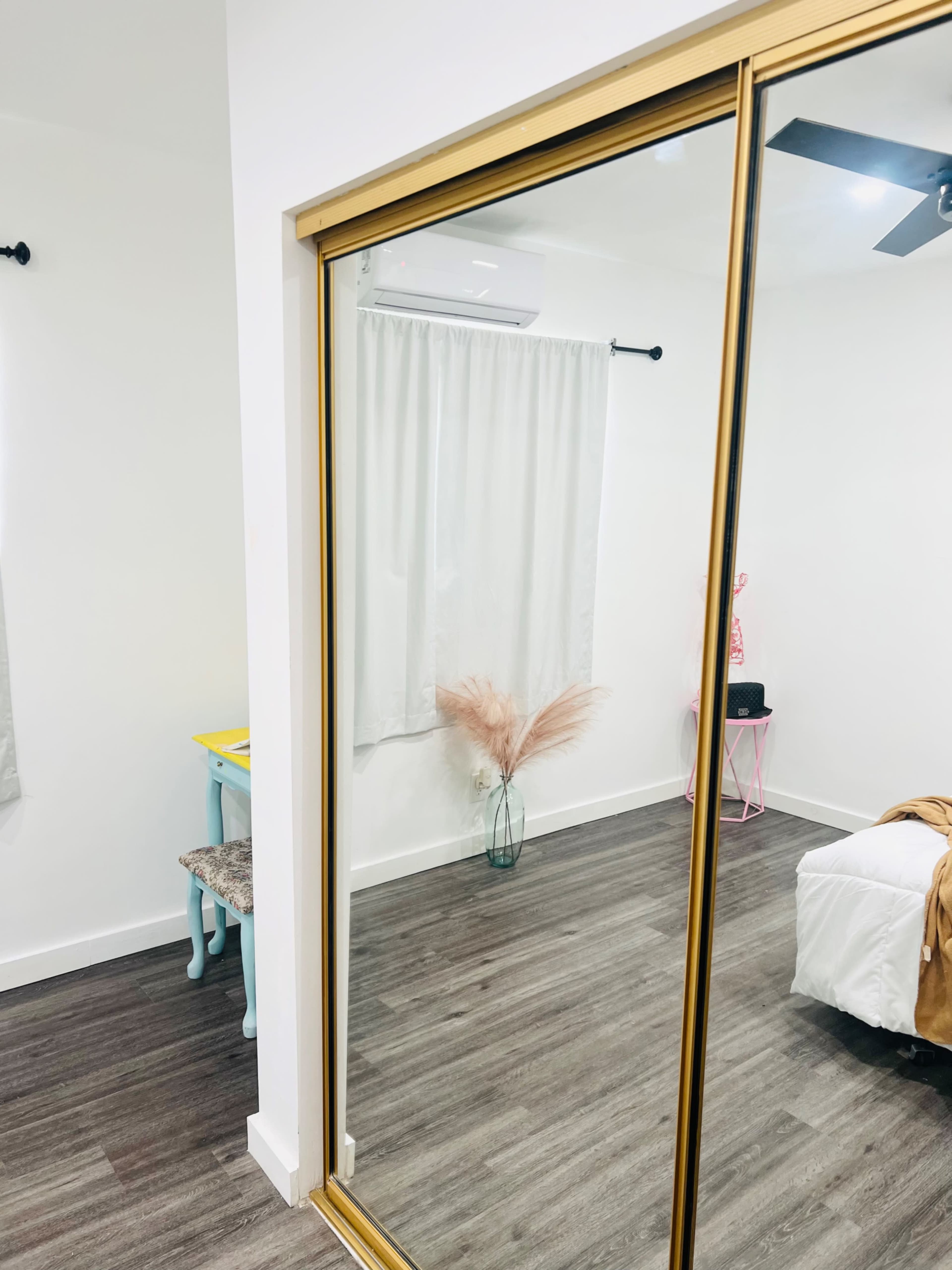 A room with a large mirrored closet door reflecting part of the space, which includes a bed, a chair, and a vase with dried plants.