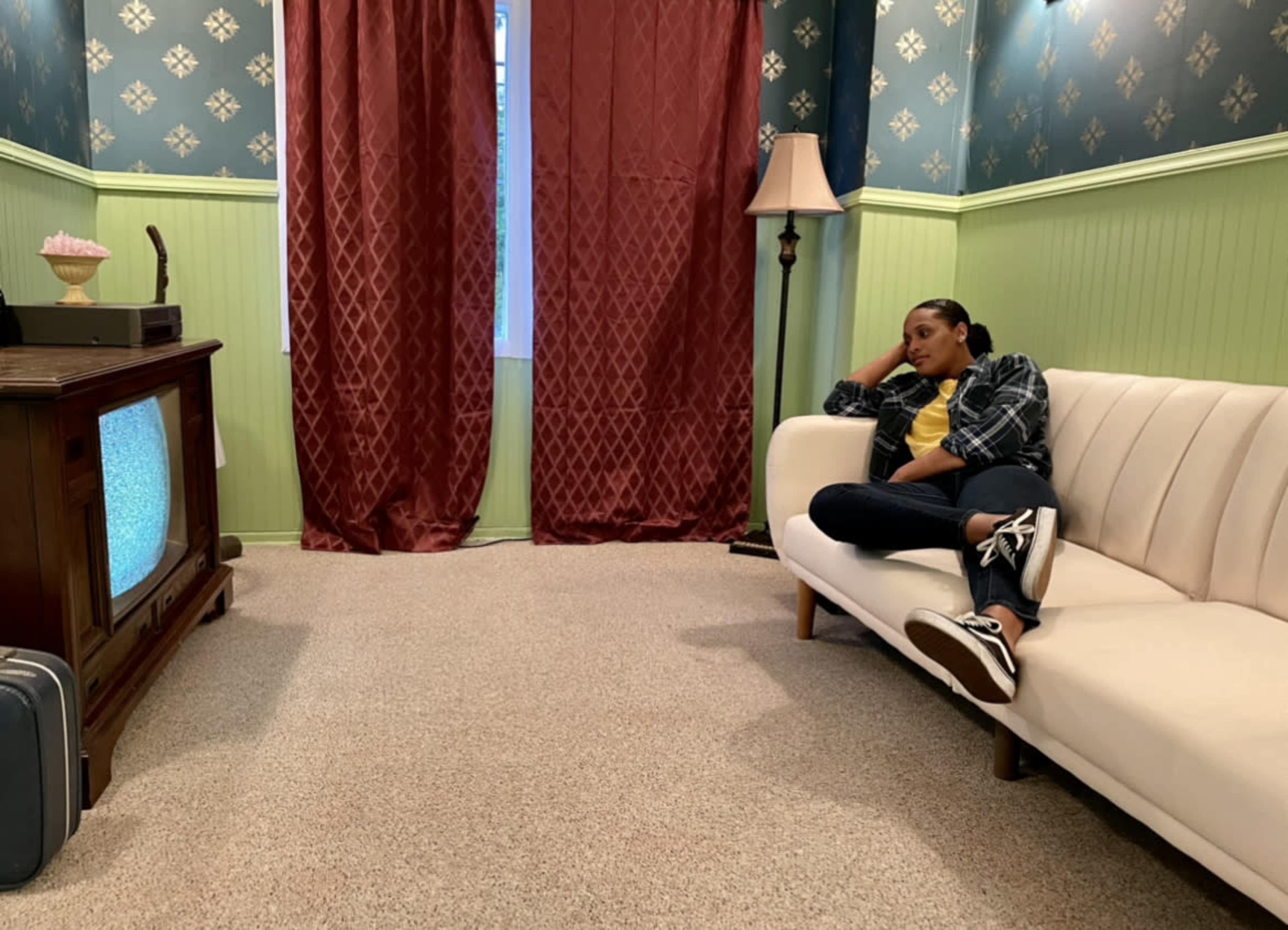 A person sits on a white couch in a room with green and blue walls, watching an old television while a lamp stands nearby.