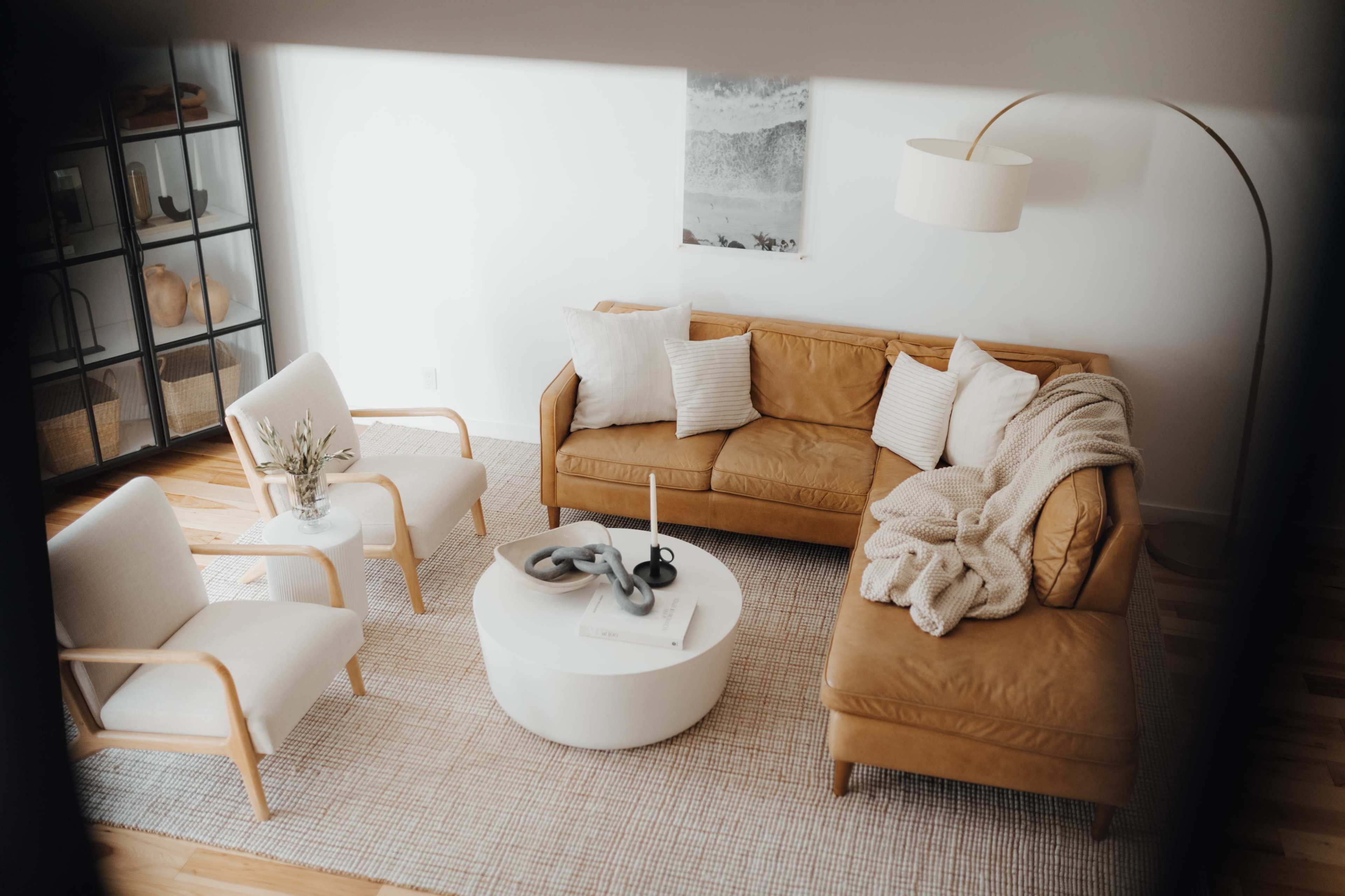 A modern living room features a tan leather sofa, two white armchairs, a round coffee table, and a textured rug.