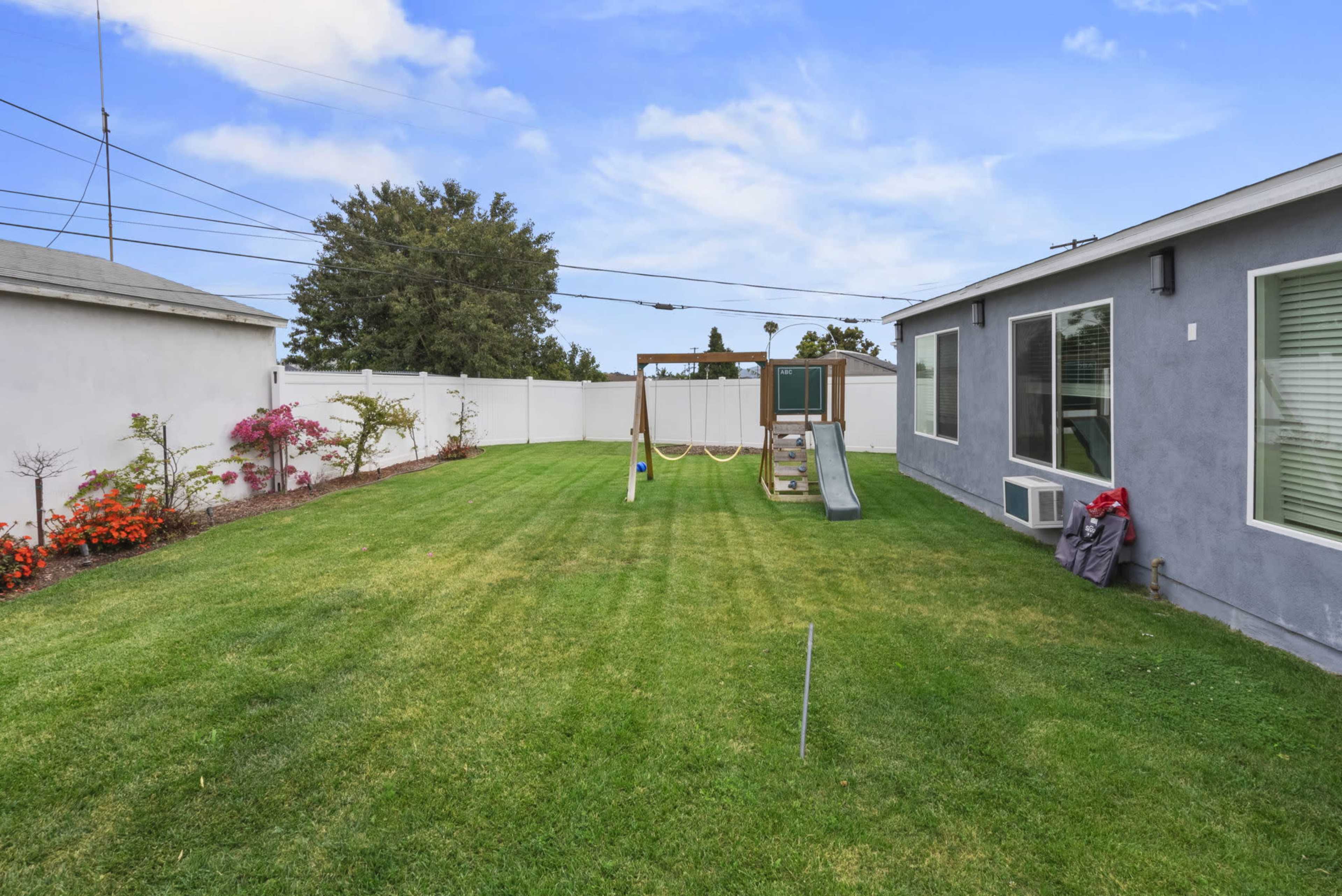 The image shows a spacious backyard with a slide and swing set, bordered by a white fence and featuring neatly manicured grass.