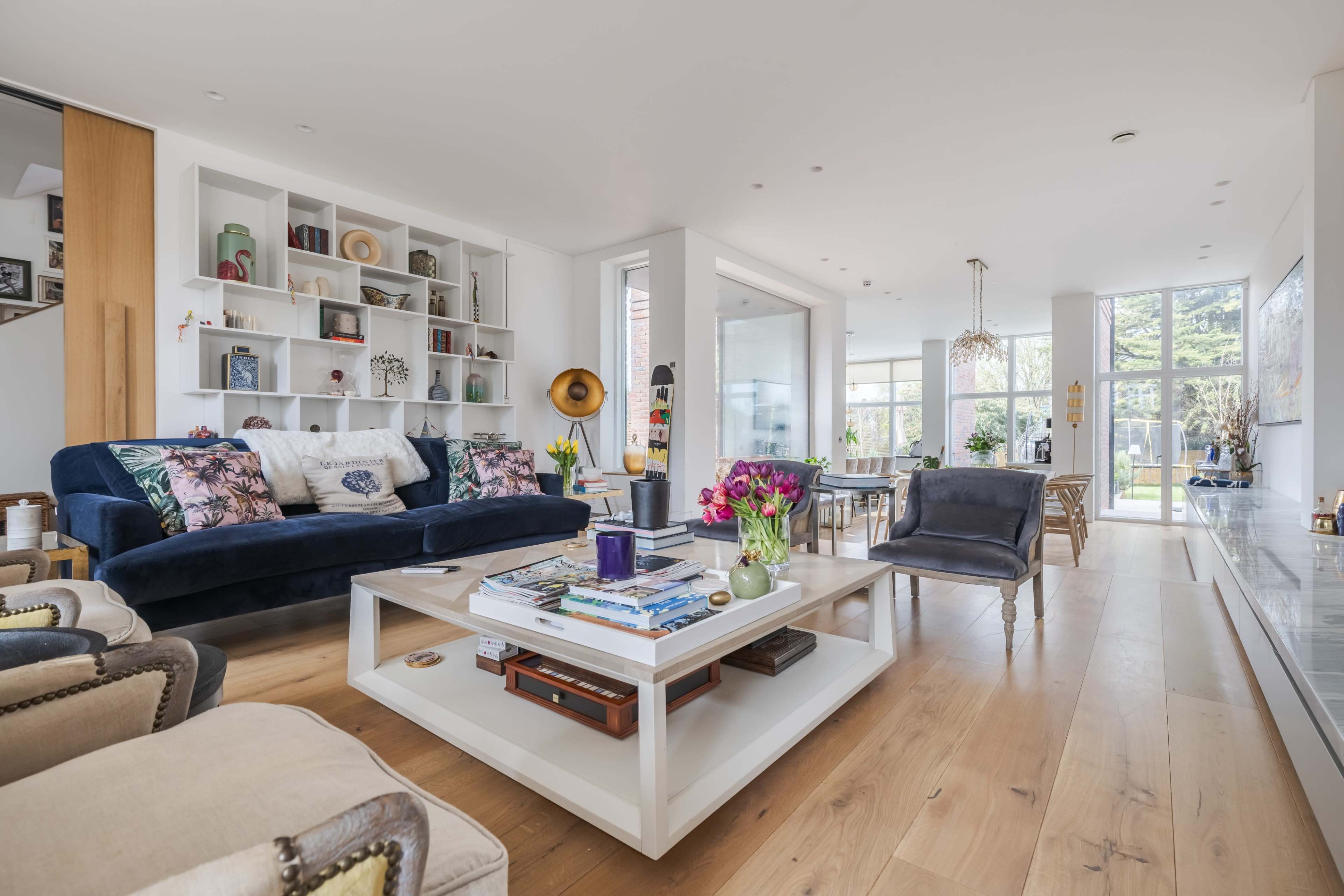 The image depicts a modern living room with a blue couch, a coffee table in the center, and a variety of decorative items on the shelves and surfaces, all set in a bright, airy space with large windows.