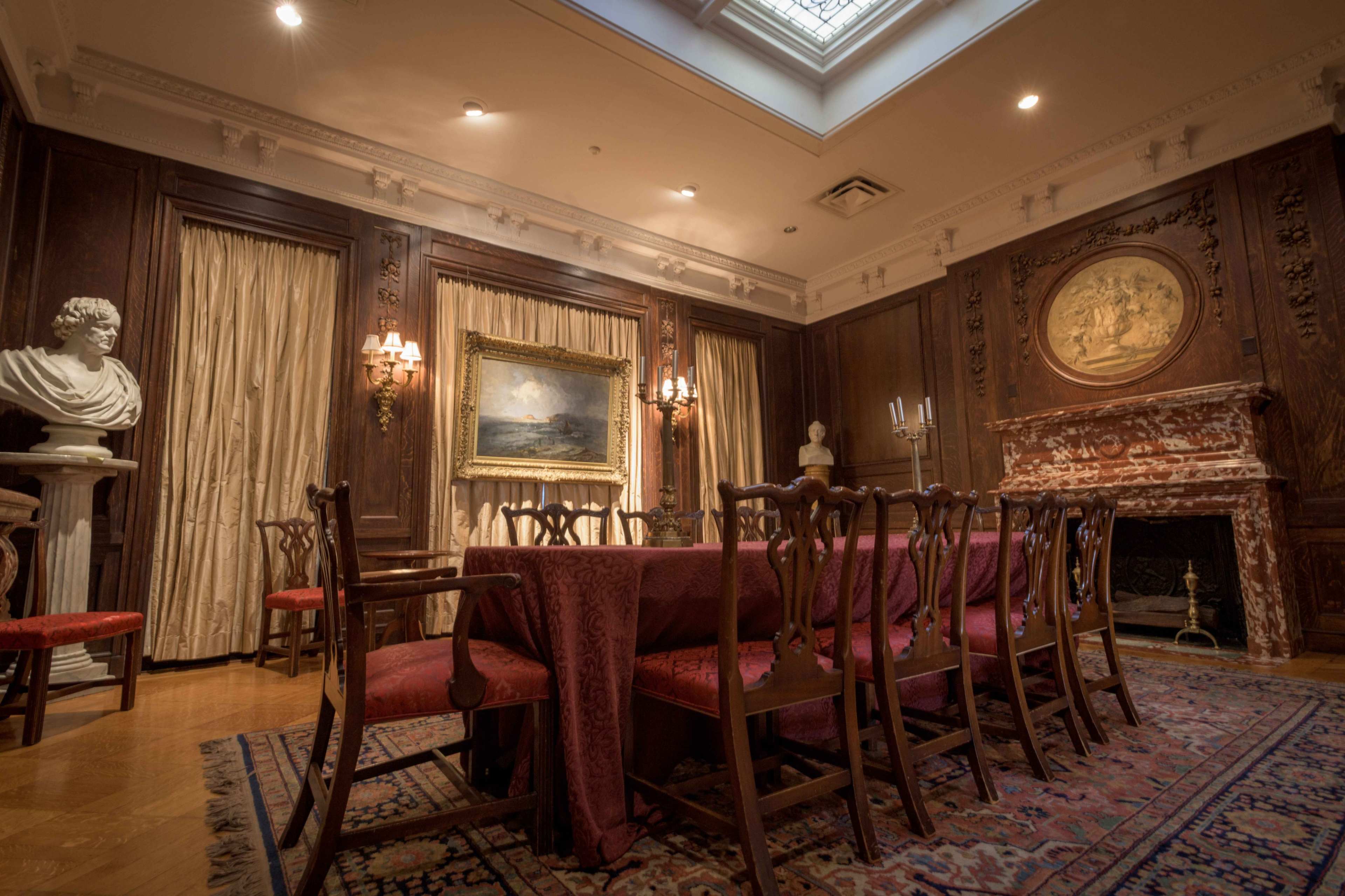 An elegant dining room featuring a long table set with red fabric, surrounded by wooden chairs, and adorned with classical decor, including a bust and a fireplace with a large circular relief.