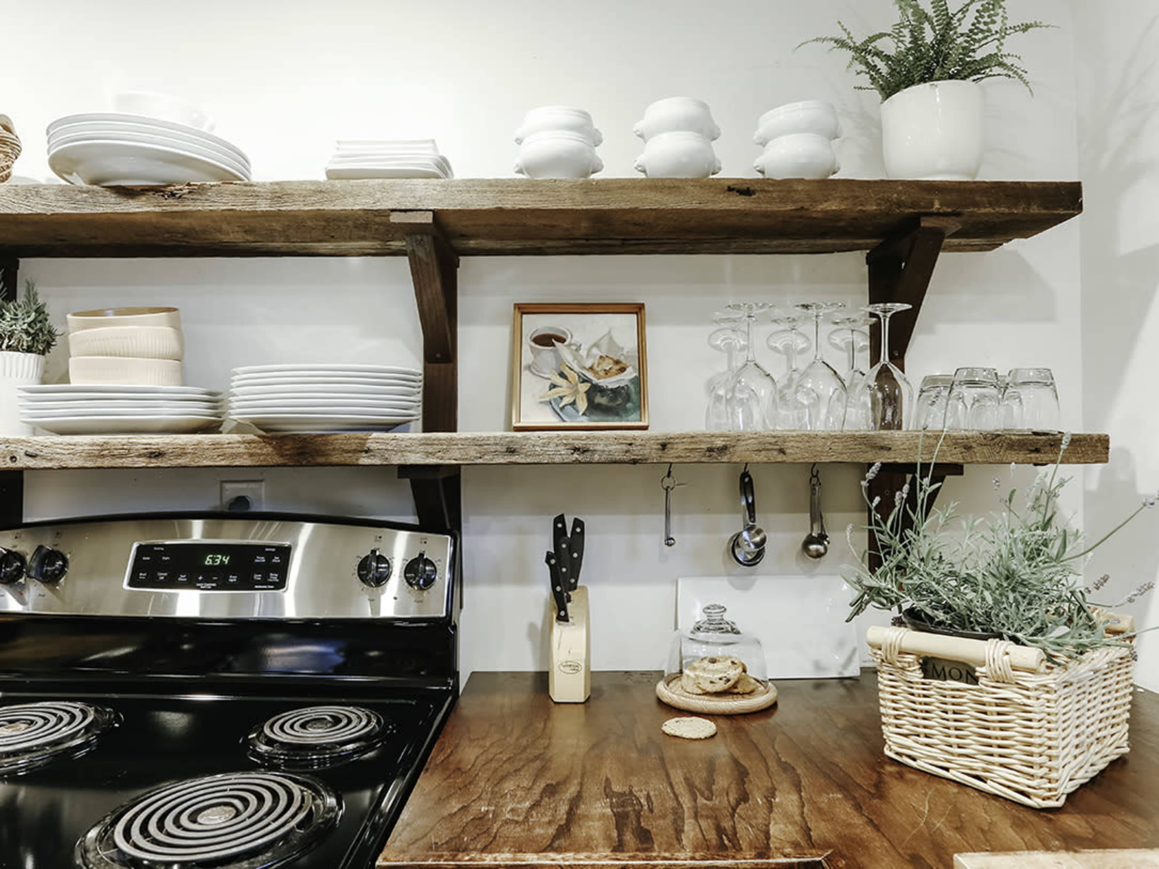 The image shows a kitchen with wooden shelves holding dishes, glassware, and decorative items, above a stove and a countertop with utensils and a biscuit on a plate.