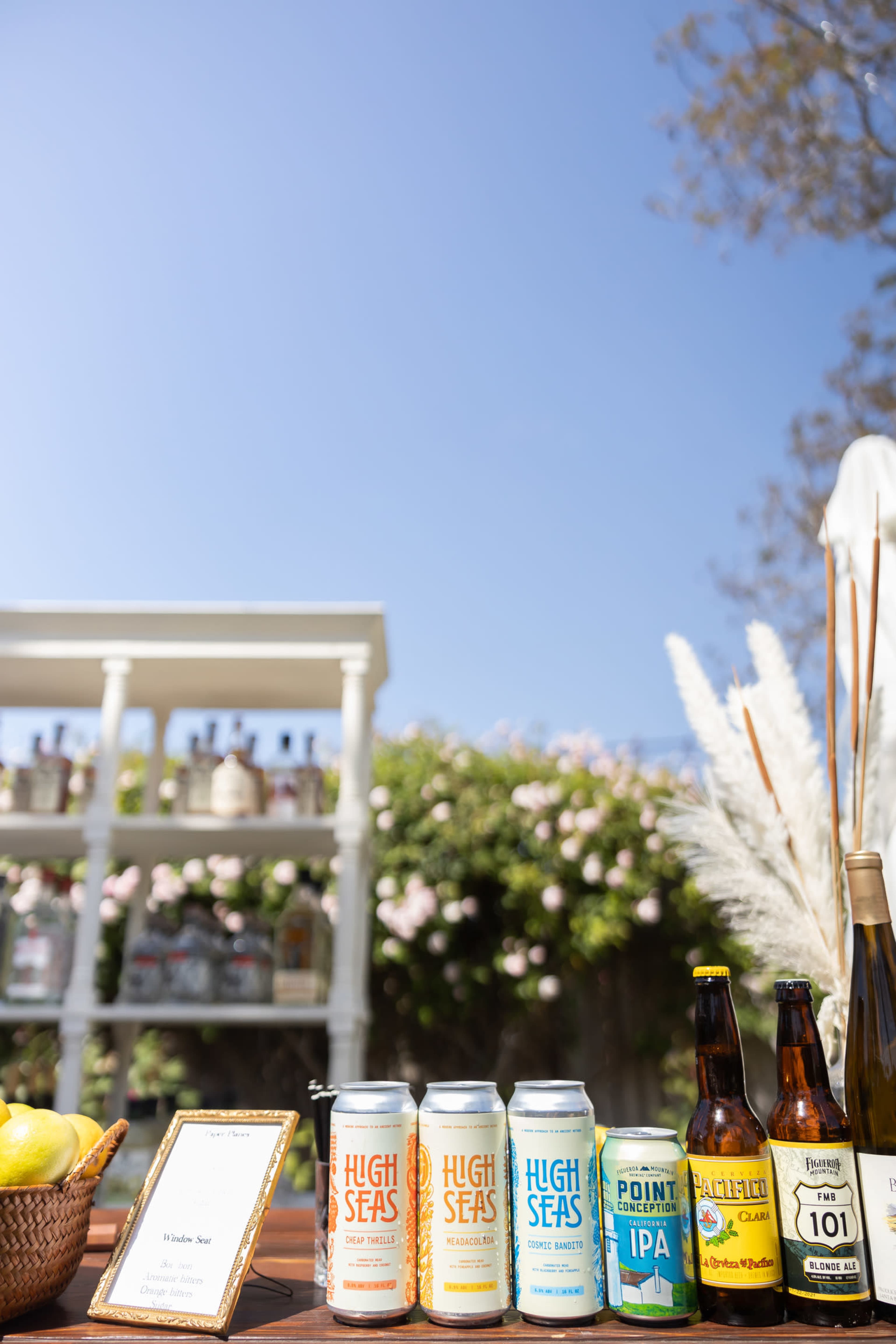 A selection of craft beers is displayed on a table under a clear blue sky, with a background of a shelf holding various bottles.