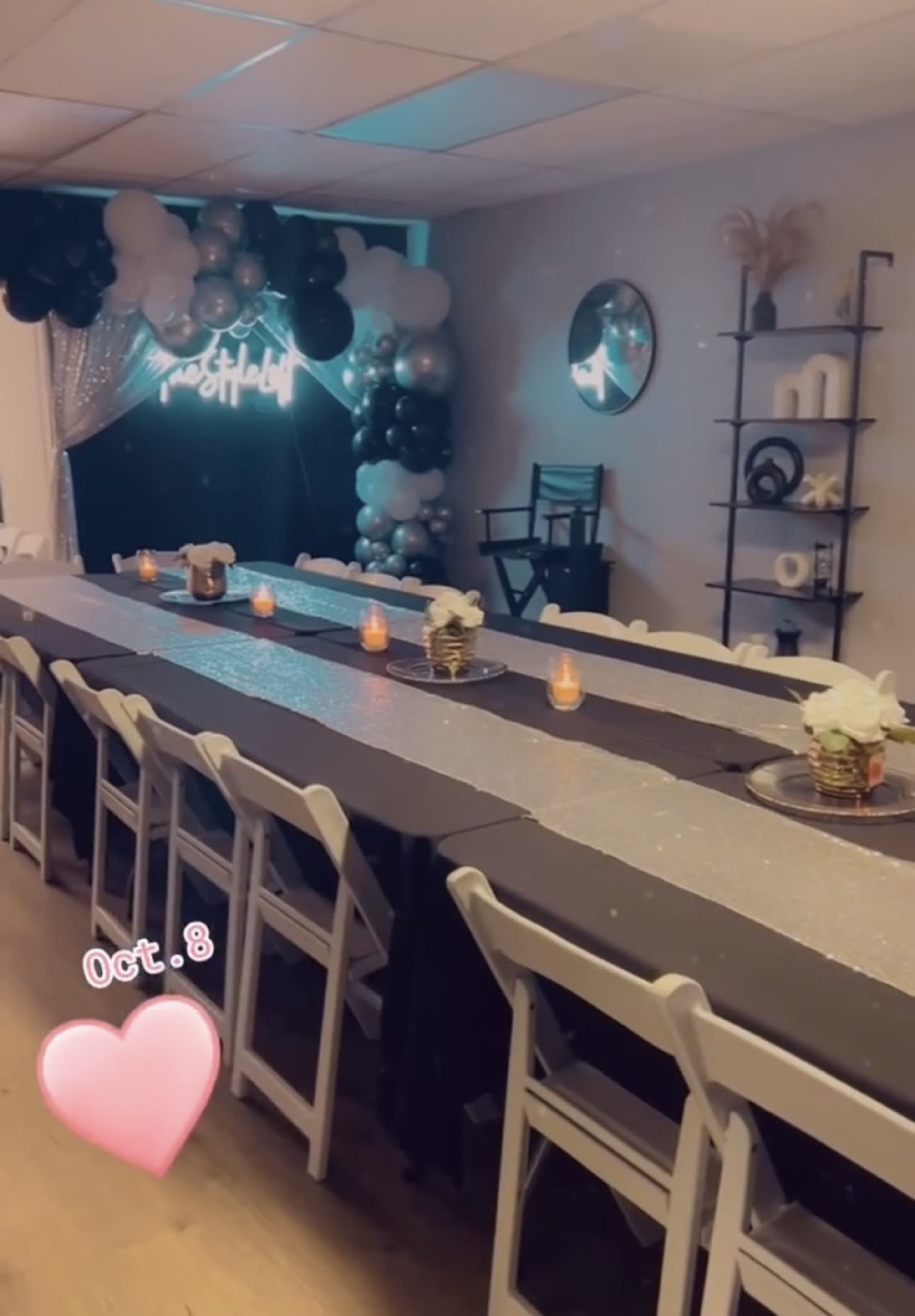 The image shows a decorated event space with a long table set for a gathering, featuring candles, floral arrangements, and a backdrop of balloons.