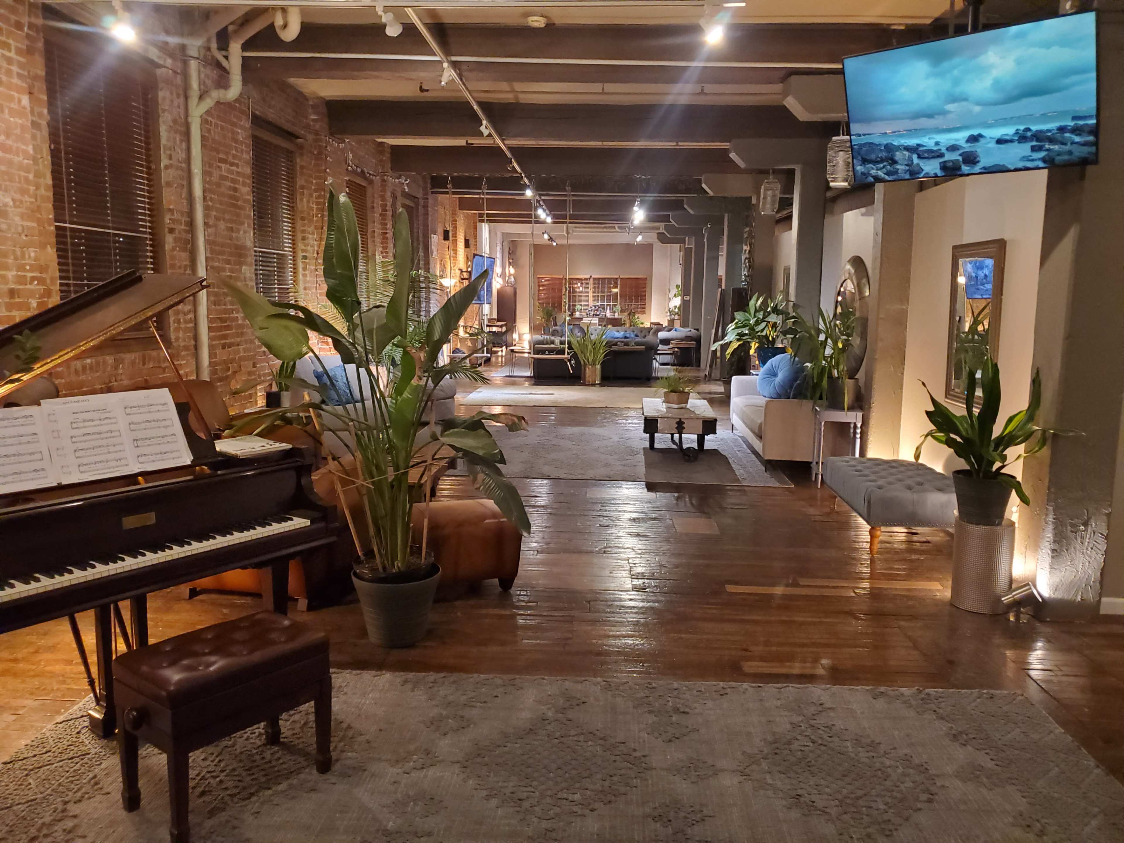 The image shows a spacious, modern interior featuring a piano, indoor plants, and a seating area illuminated by soft lighting.