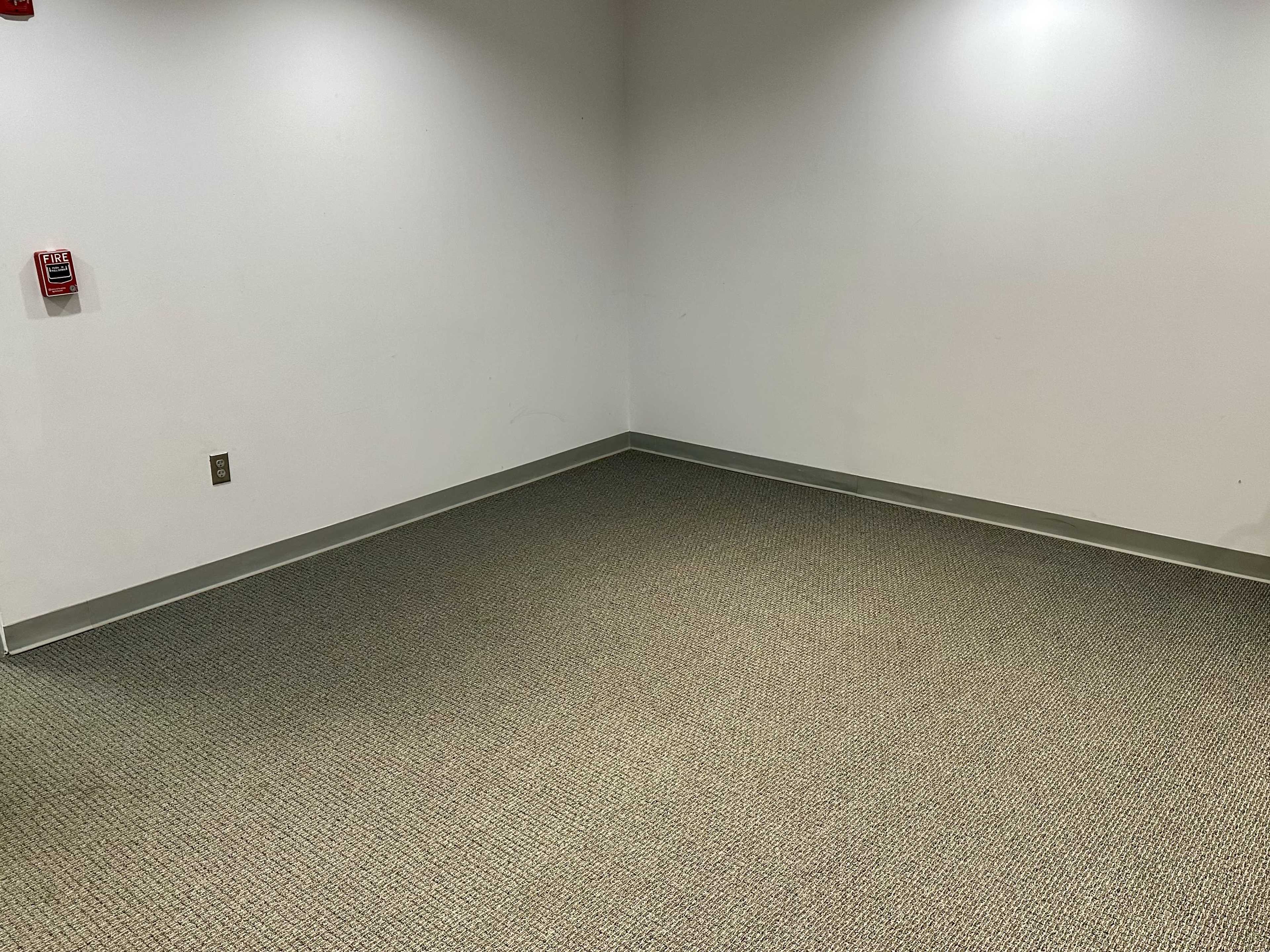 The image shows a sparsely furnished room with gray carpet, white walls, and minimal lighting.