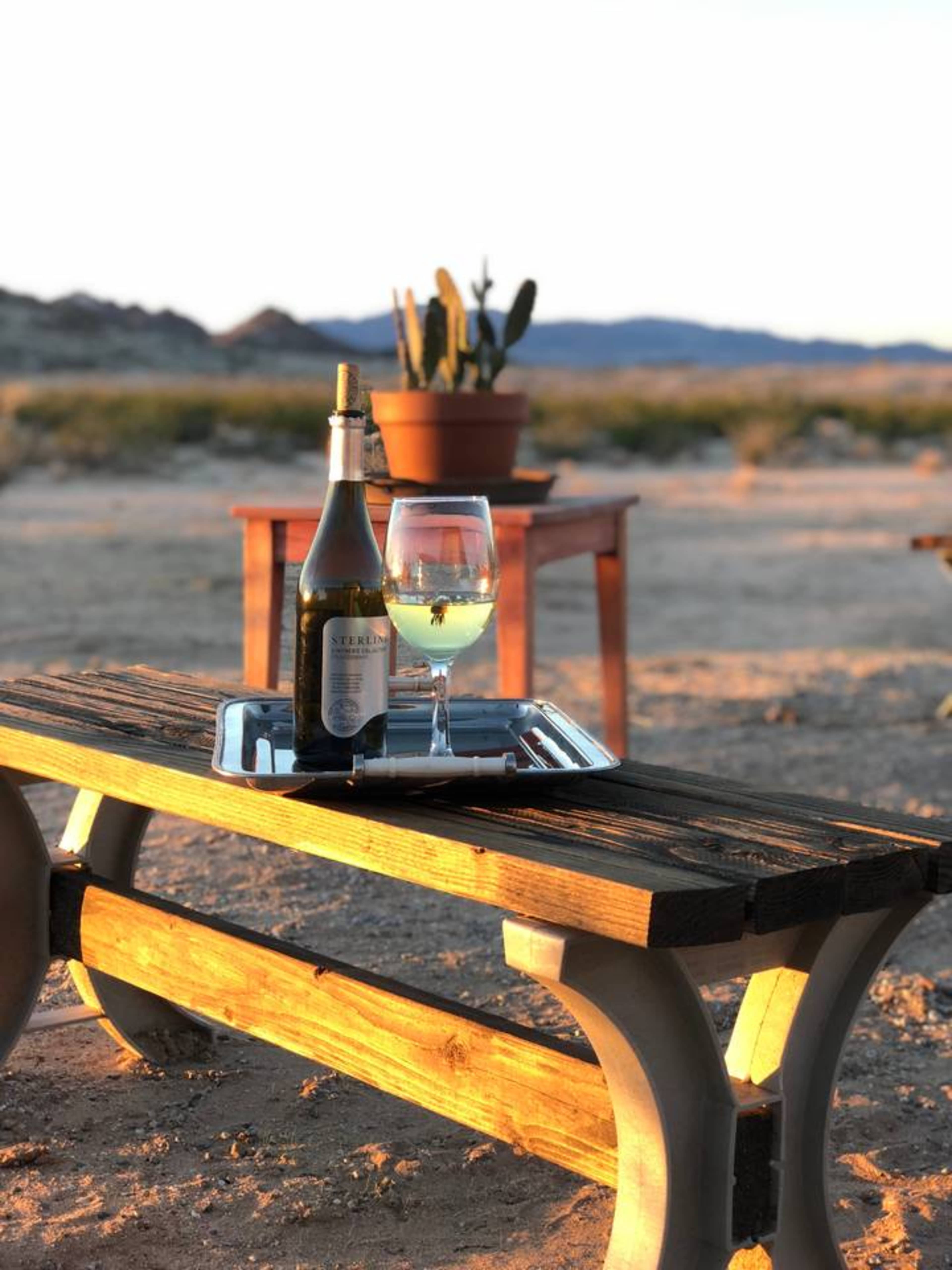 A bottle of wine and a glass sit on a wooden table with a cactus in the background against a desert landscape at sunset.
