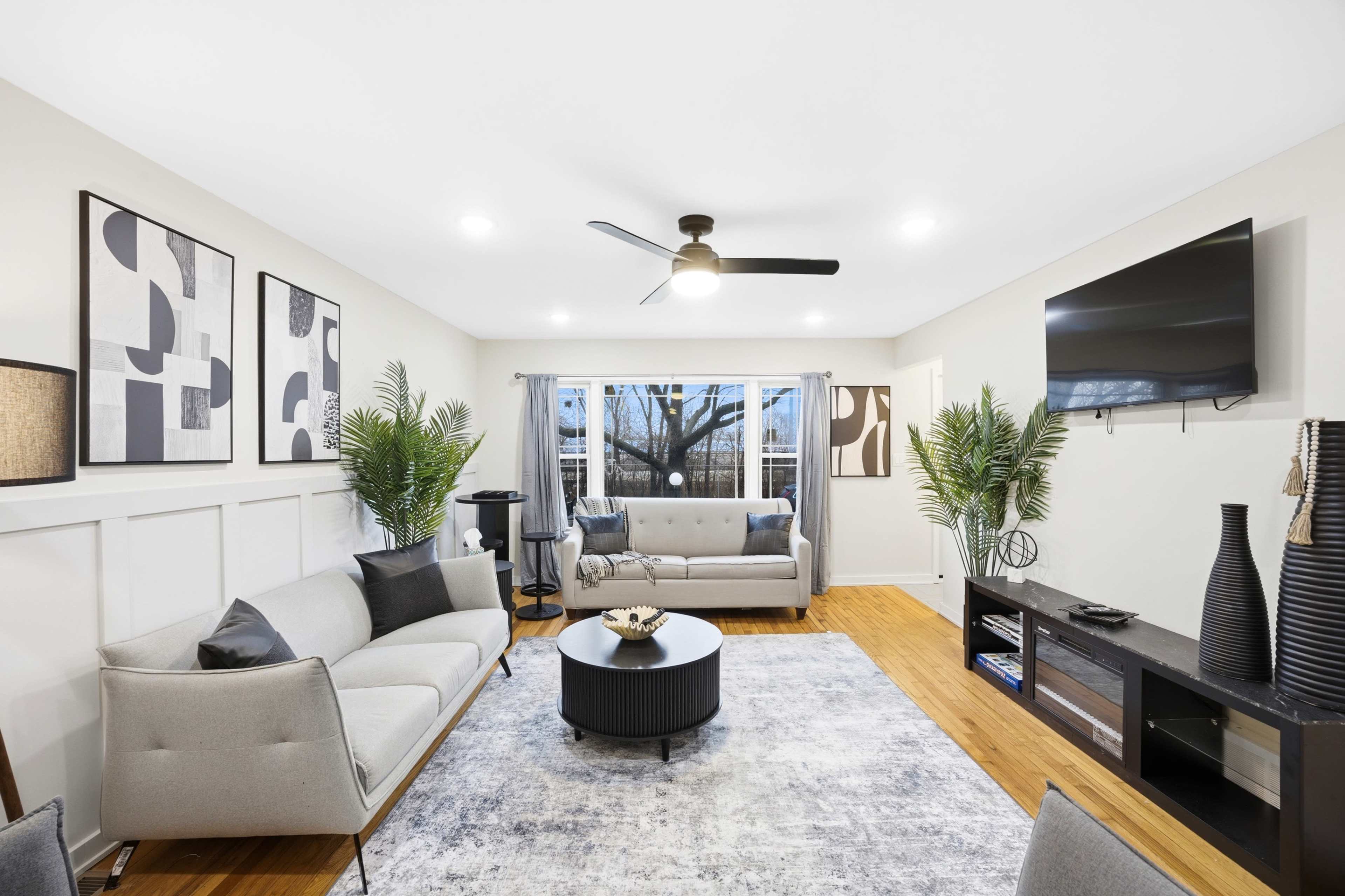 The image shows a modern living room with a neutral color palette, featuring two sofas, a round coffee table, a television mounted on the wall, and decorative plants.