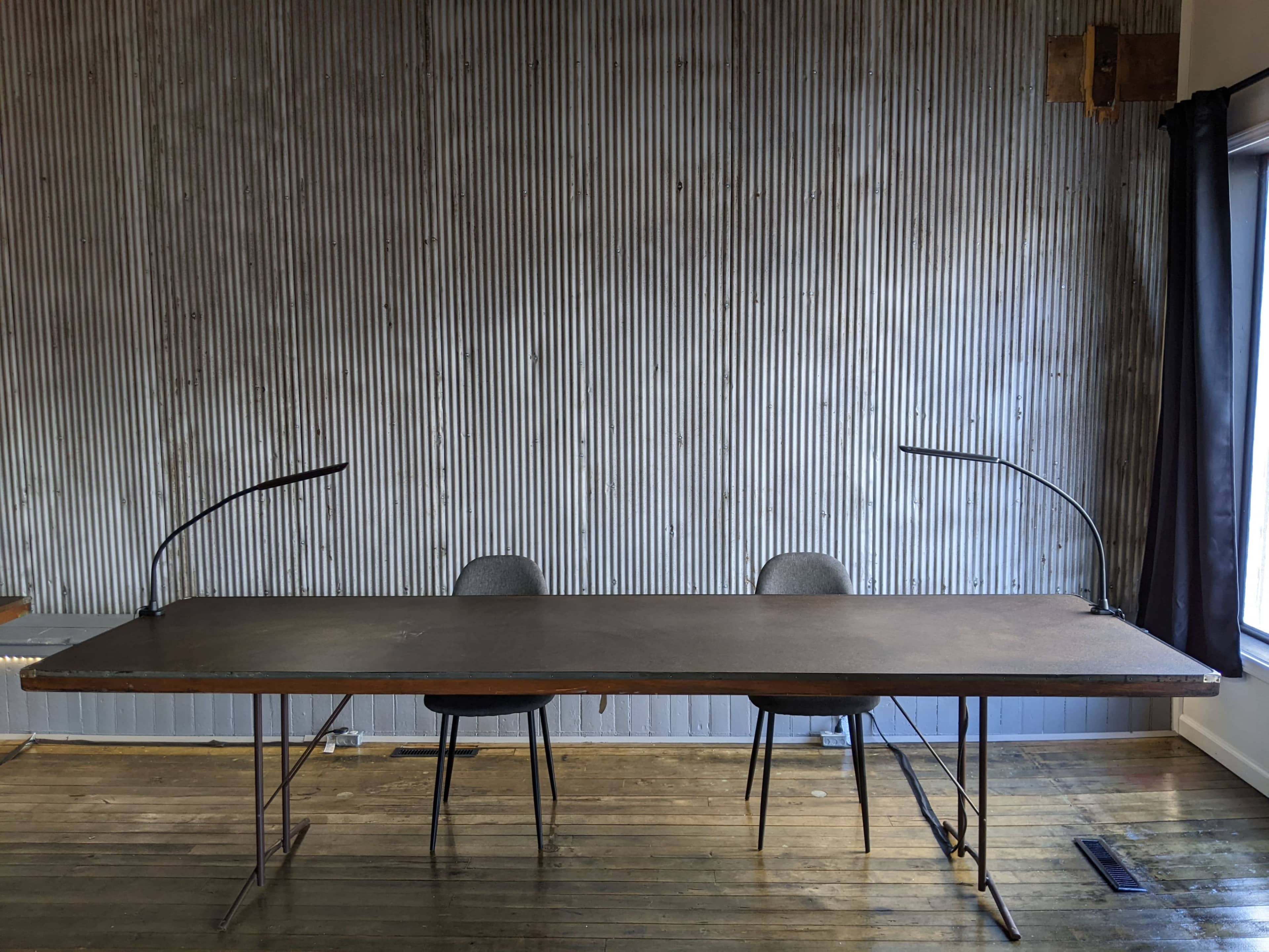 The image shows a long wooden table with two chairs on either side, placed against a corrugated metal wall.