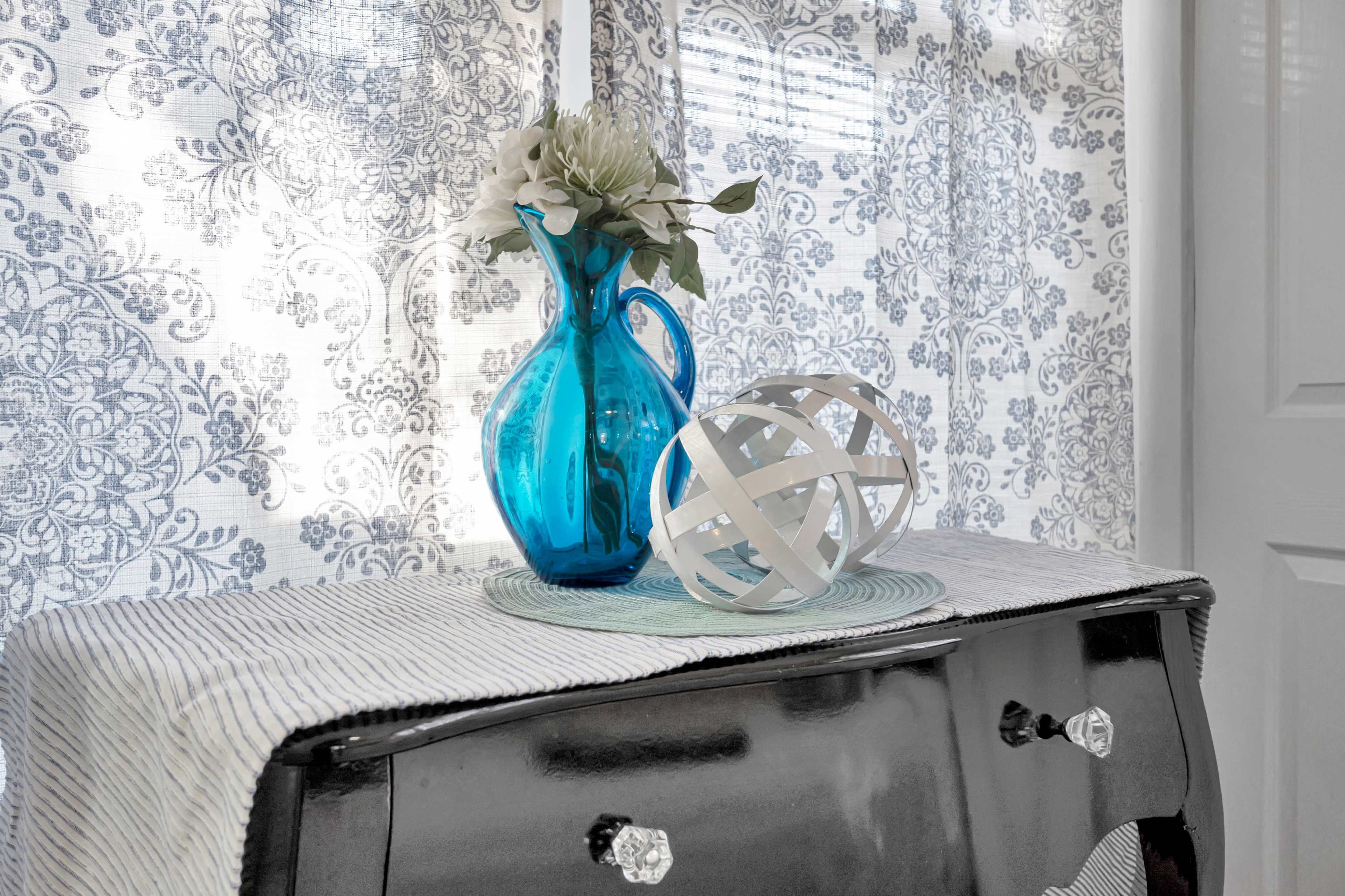 A blue glass pitcher with a floral arrangement and a white decorative orb sit atop a black dresser covered with a striped cloth.