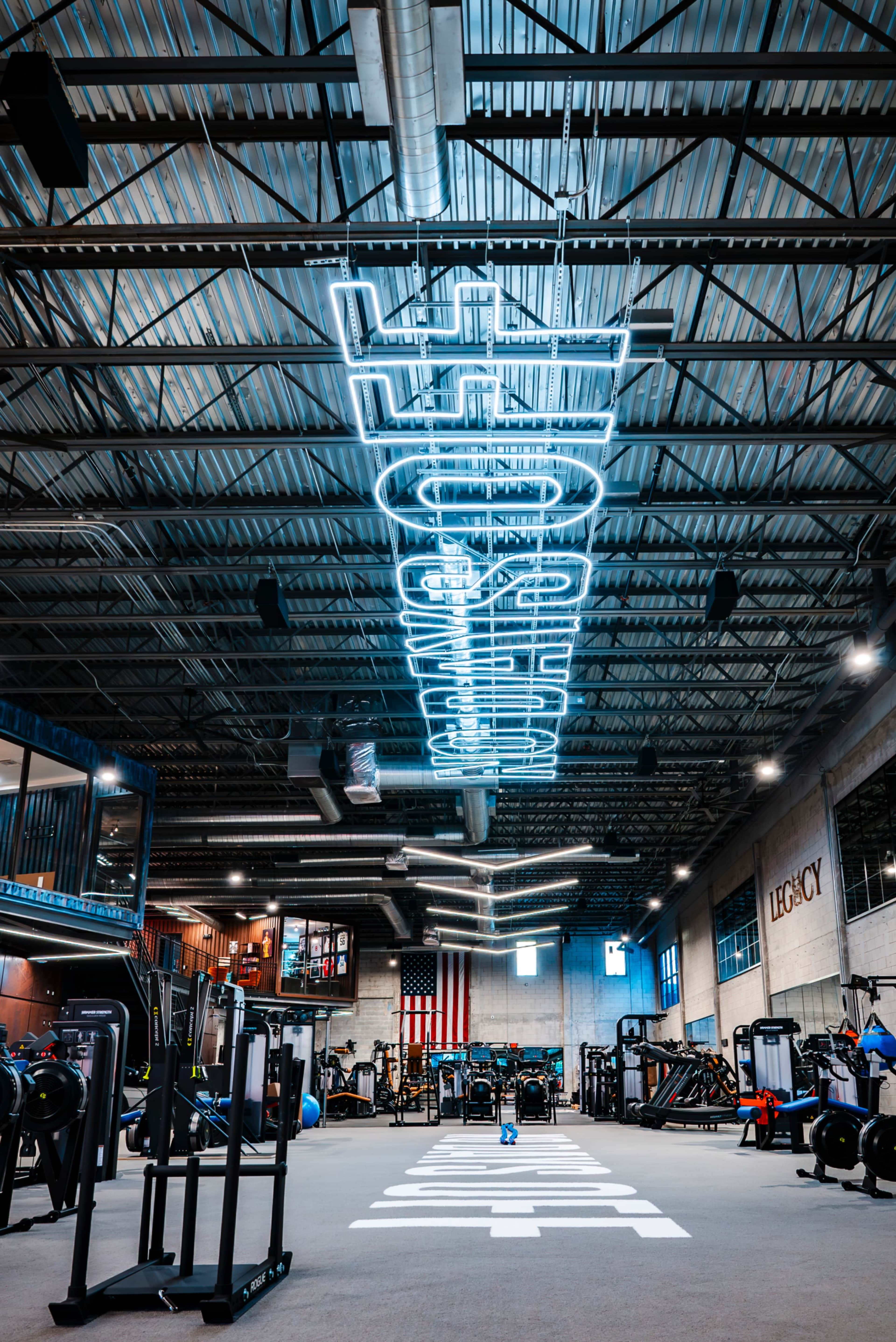 The image shows a gym interior featuring workout equipment, a neon sign hanging from the ceiling, and an American flag on the wall.