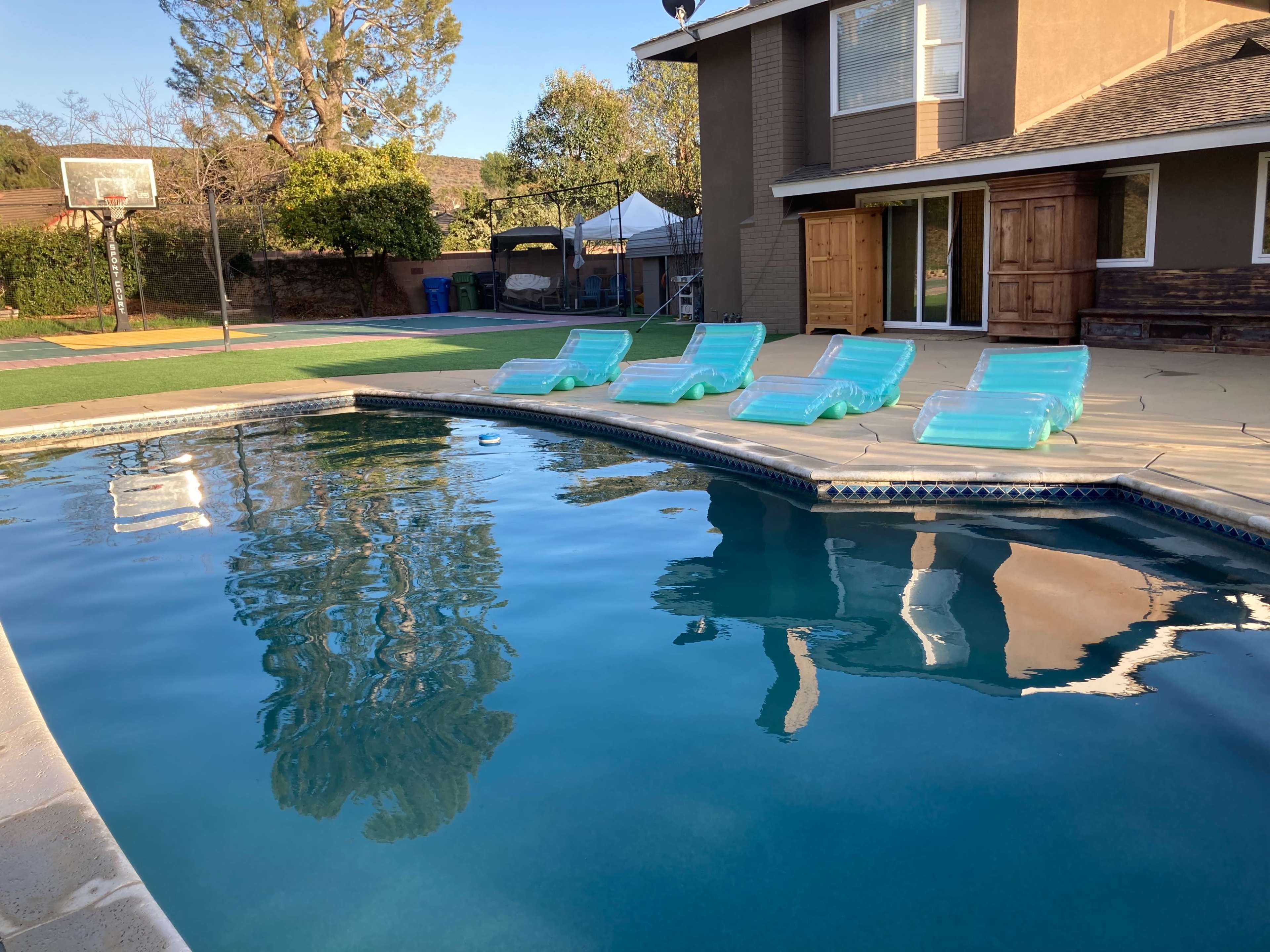 A swimming pool with five turquoise lounge chairs arranged neatly around it, reflecting the surrounding trees and a house with large windows.
