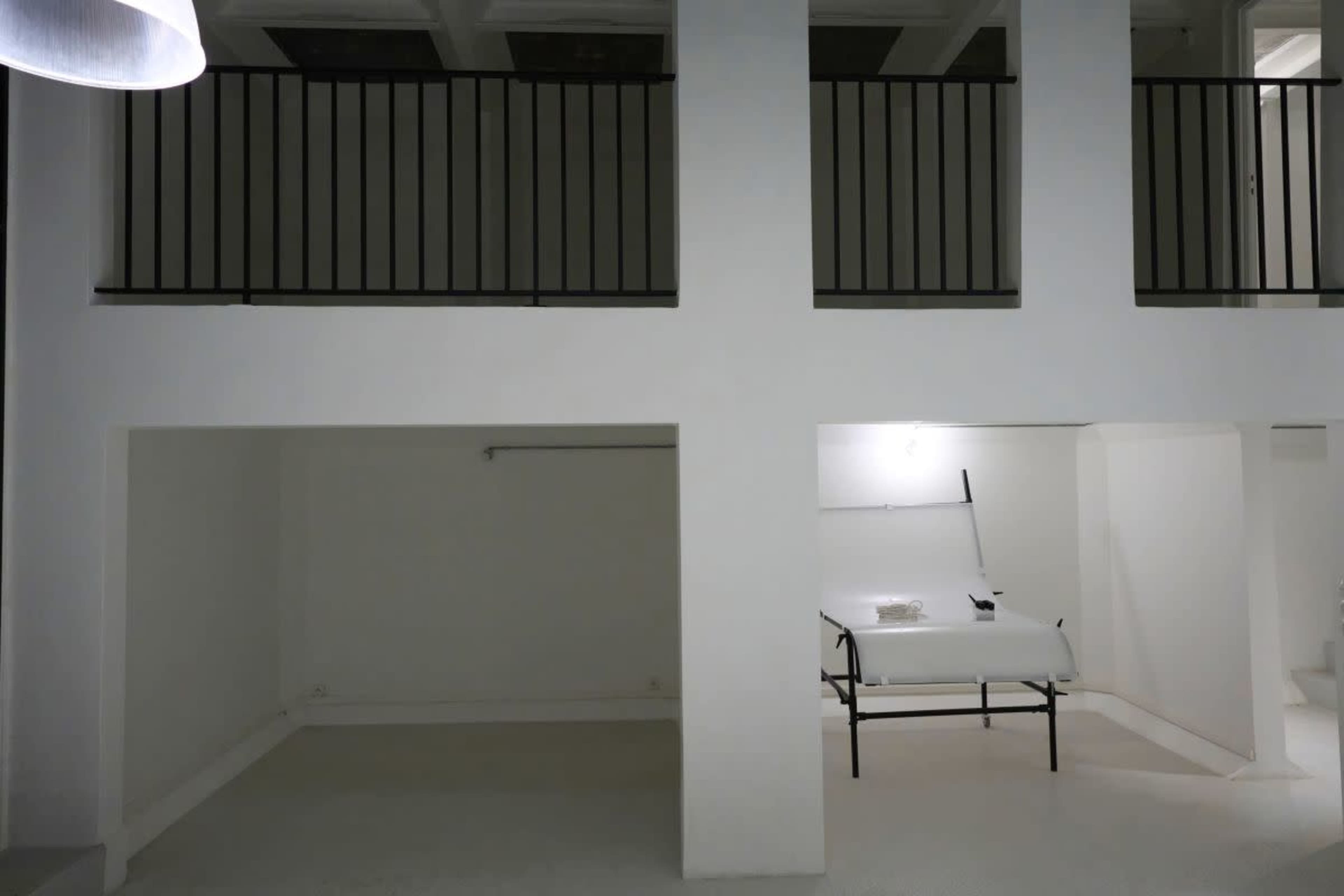 The image shows a minimalistic, well-lit interior space with white walls and a table in the corner, featuring a black railing on the upper level.
