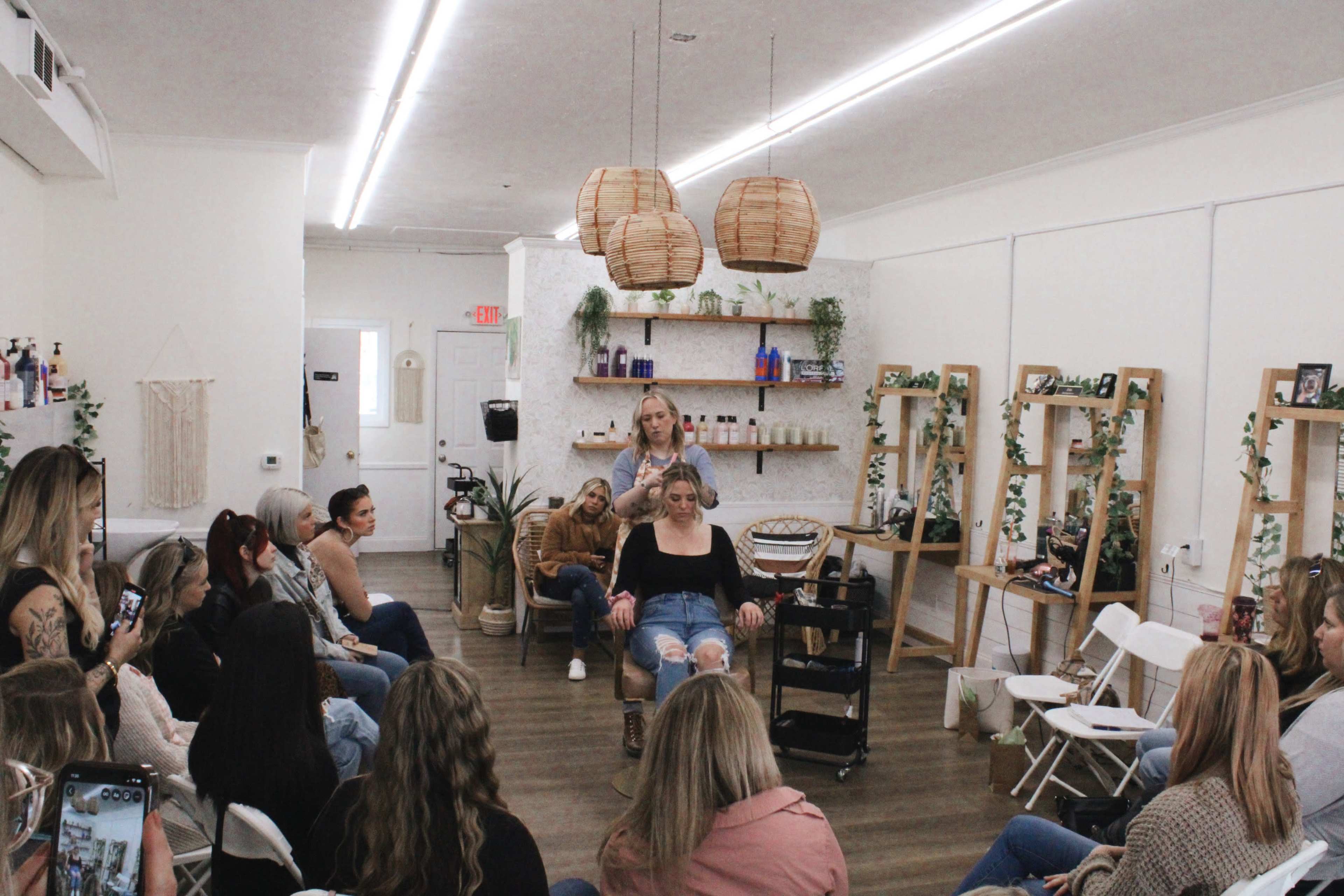 A group of people sits in a salon, watching a stylist demonstrate hair techniques on a model in a chair.