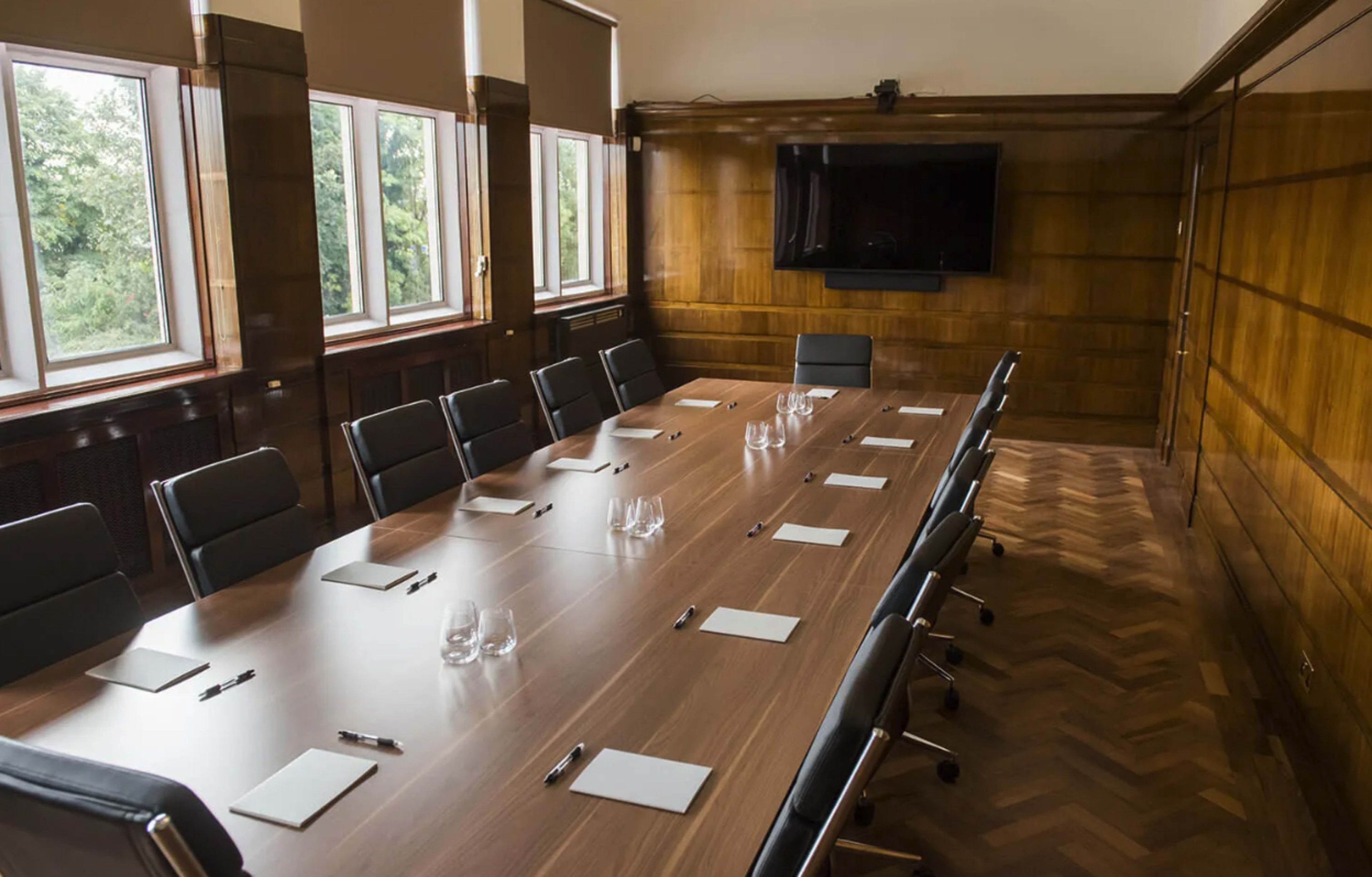 A large conference room features a wooden table surrounded by black chairs, with a television on the wall and notepads set at each place.