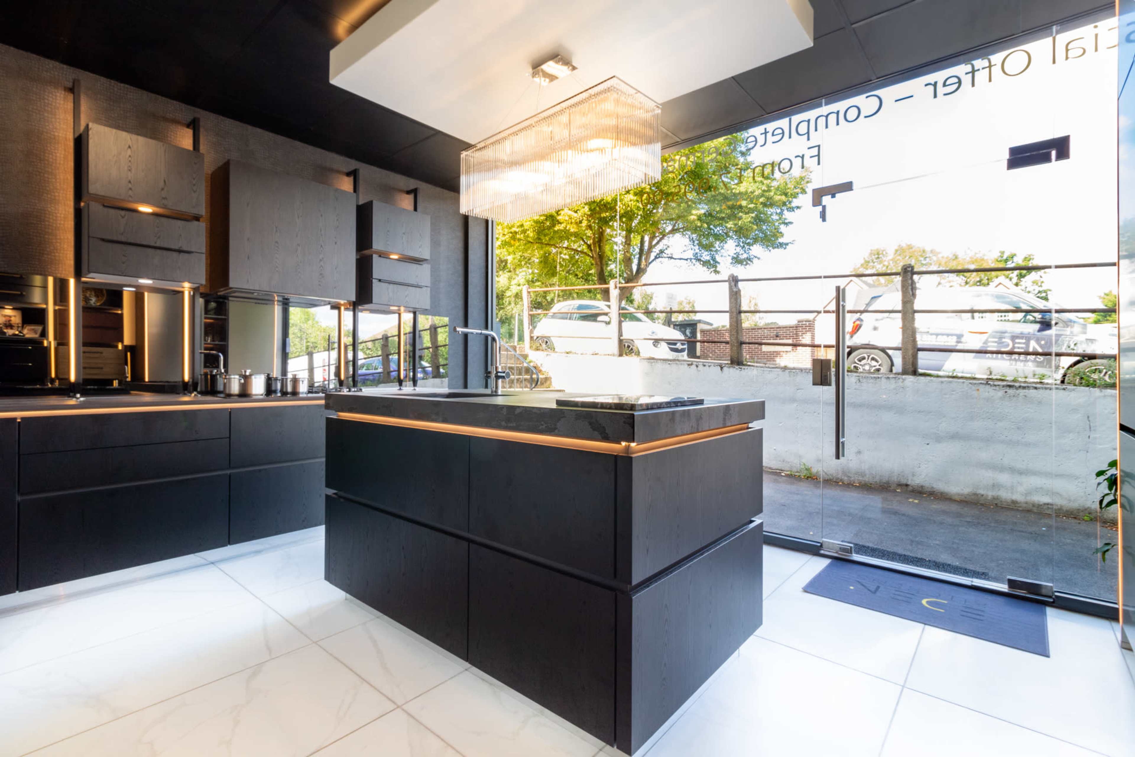 The image shows a modern kitchen interior with a central island, dark cabinetry, a chandelier, and a view of cars parked outside through large glass windows.