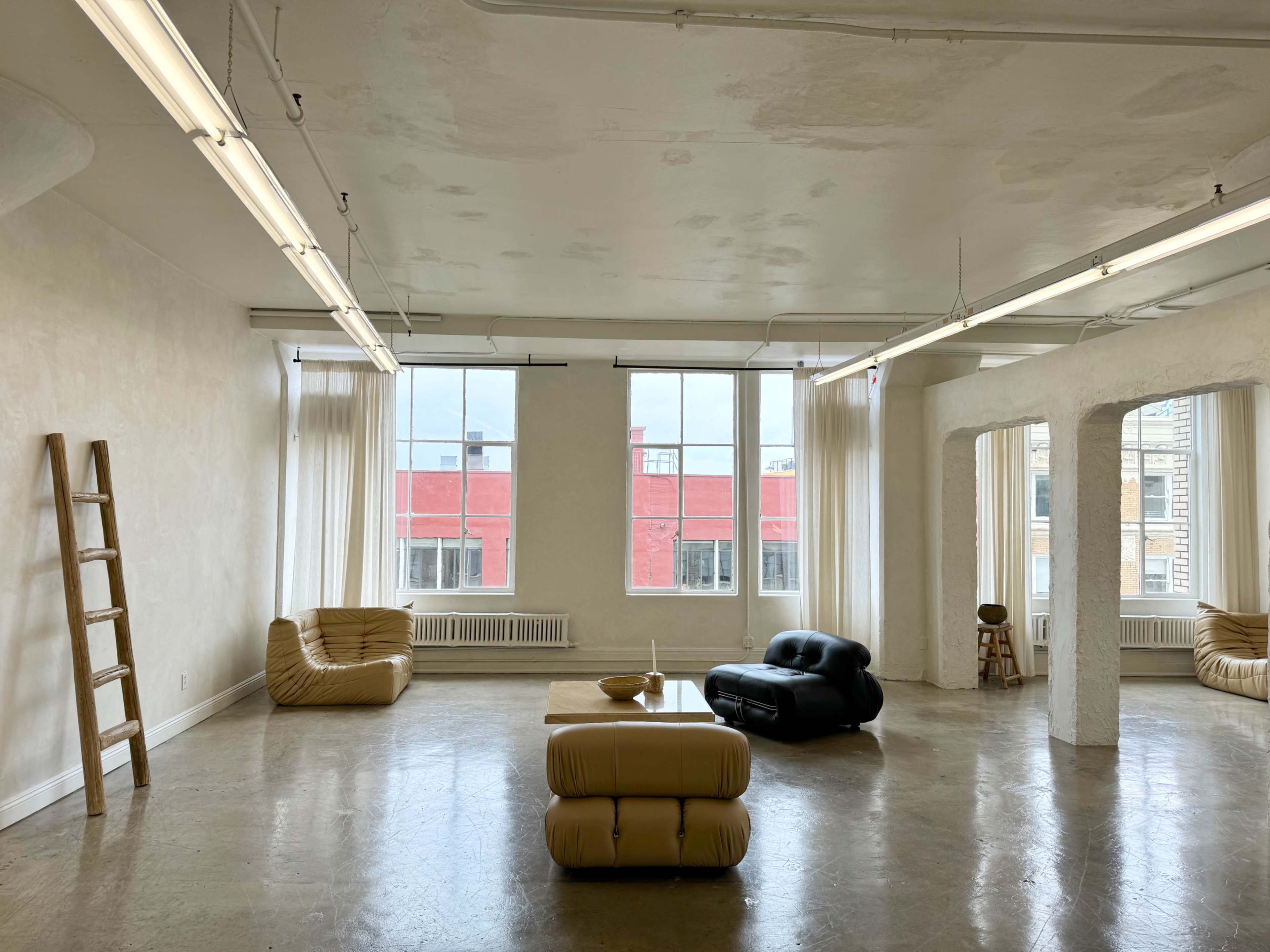 A spacious, well-lit room with large windows, minimalist furniture, and a polished concrete floor.