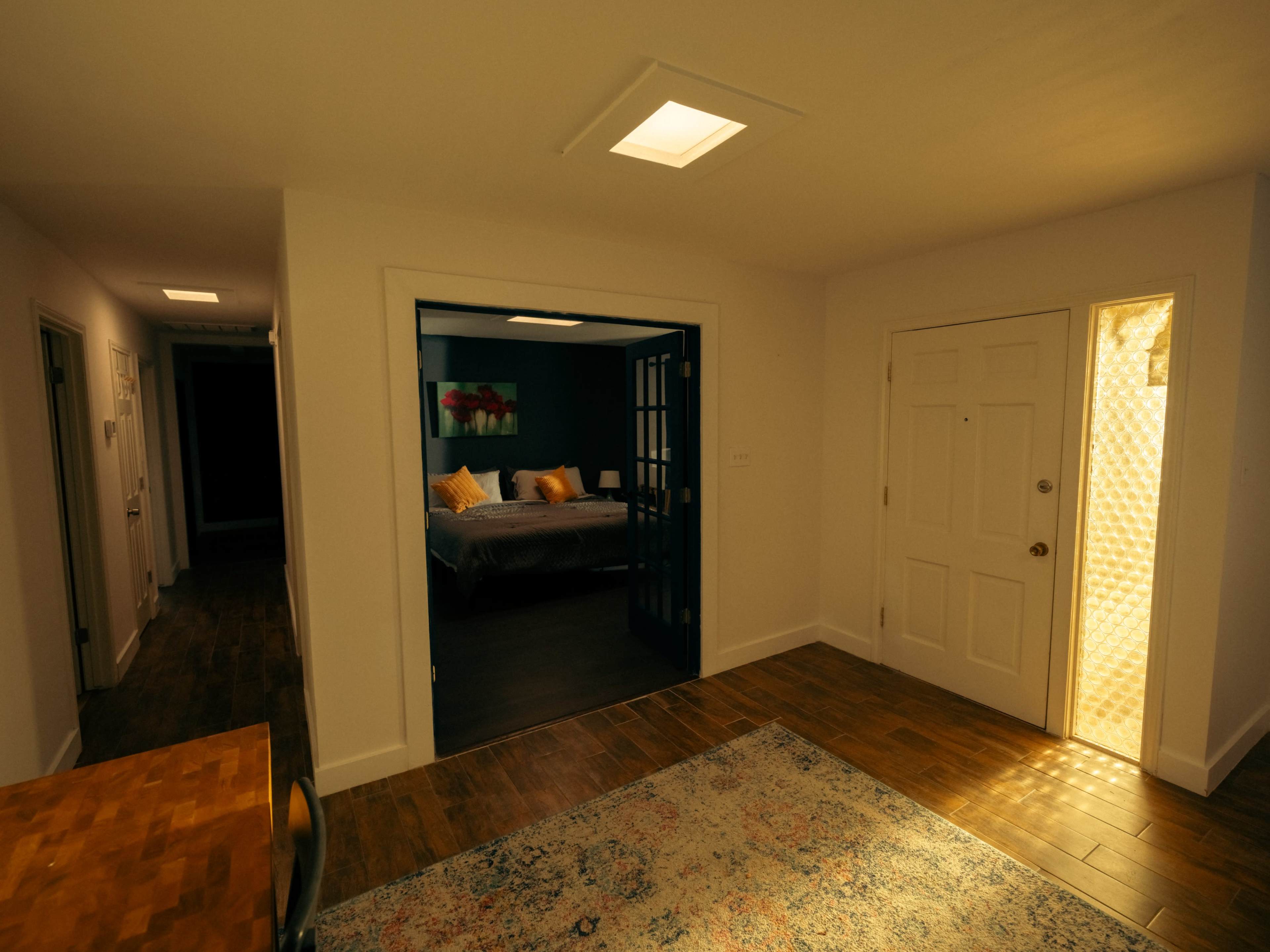 The image shows a dimly lit, modern entryway that leads to a bedroom, with a door on the right and a blue-framed doorway on the left.