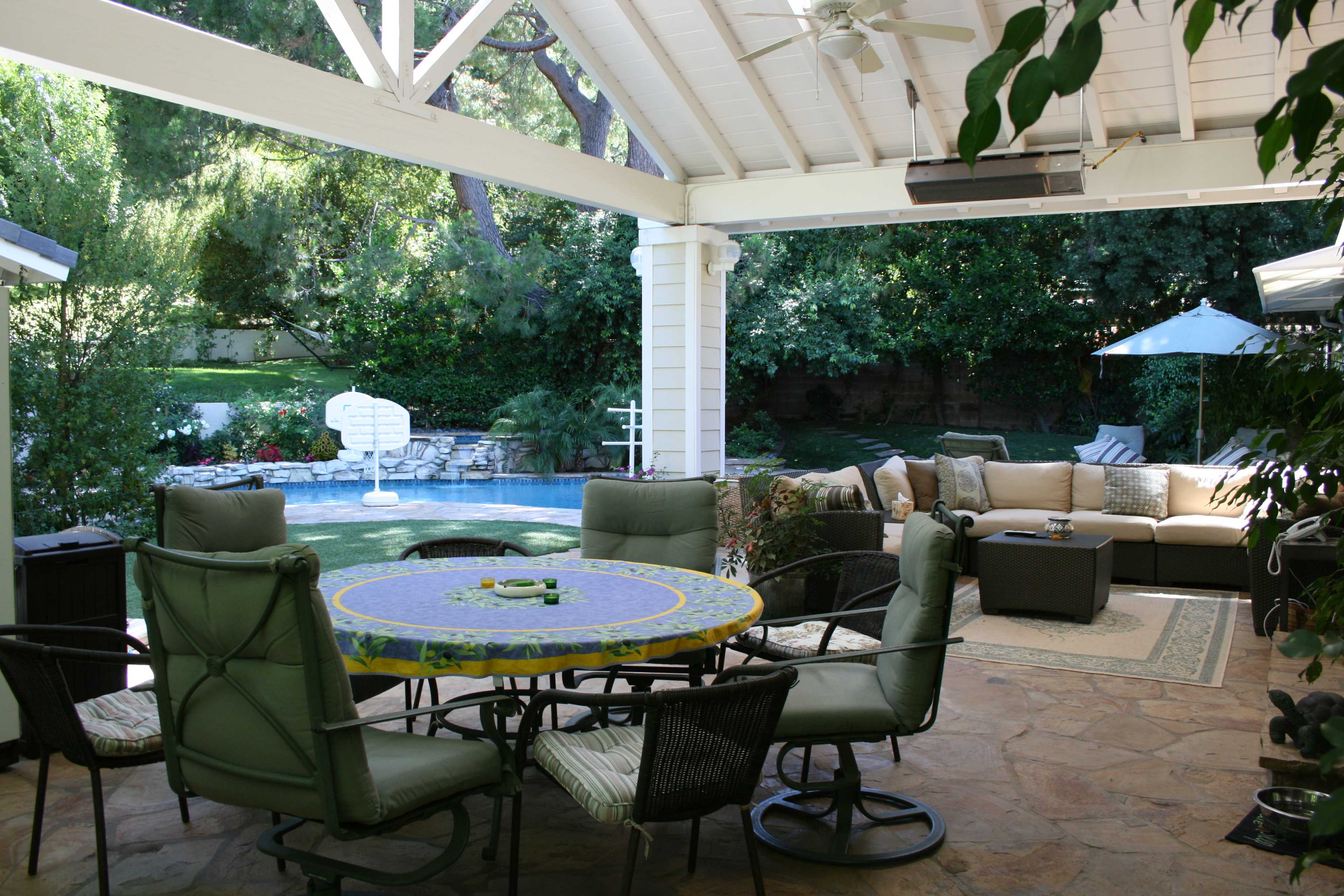 A backyard patio with a round dining table, green chairs, and a seating area beside a pool surrounded by greenery.