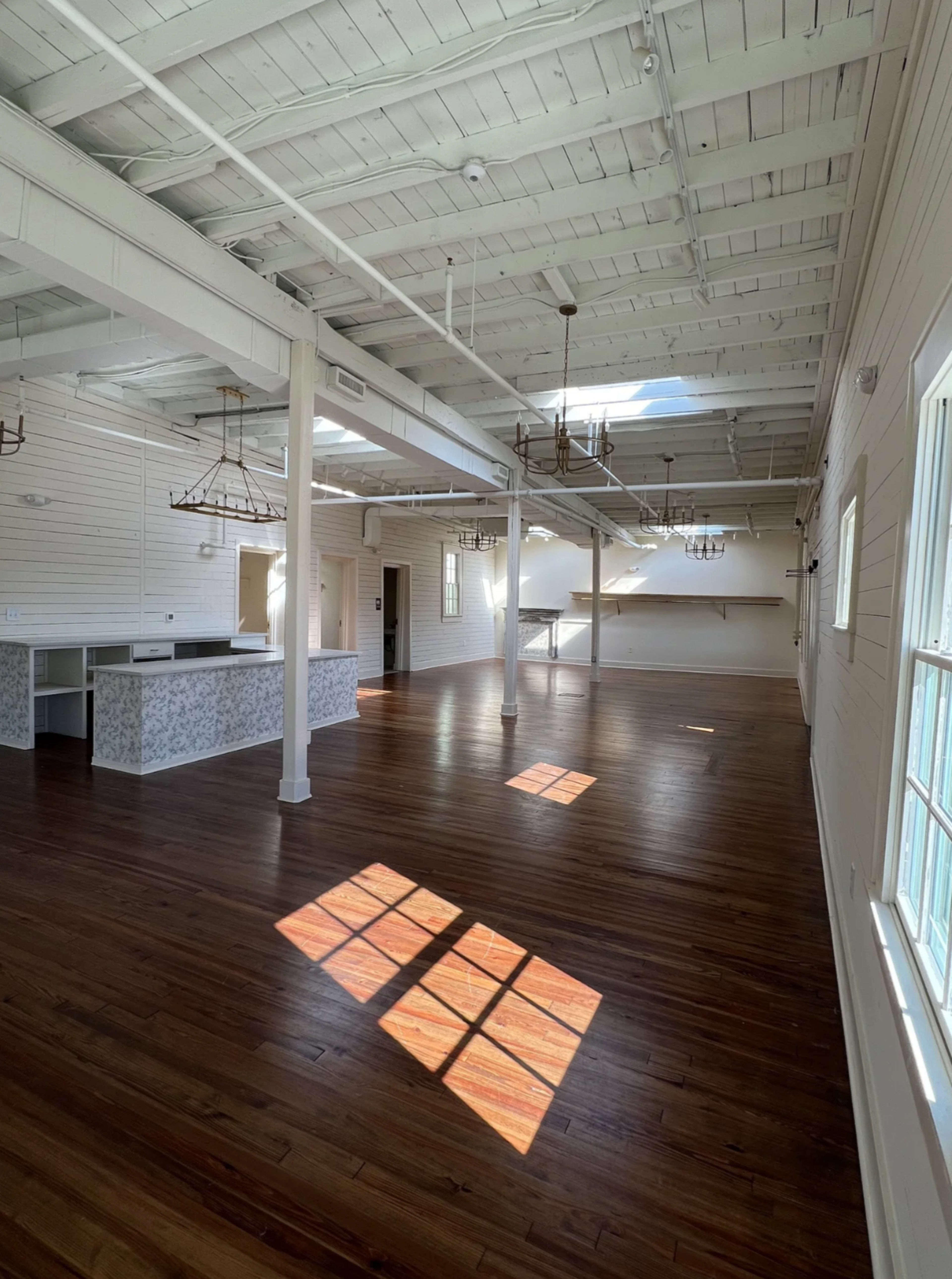The image shows a spacious interior with high ceilings, wooden floors, and multiple windows allowing natural light to illuminate the room.