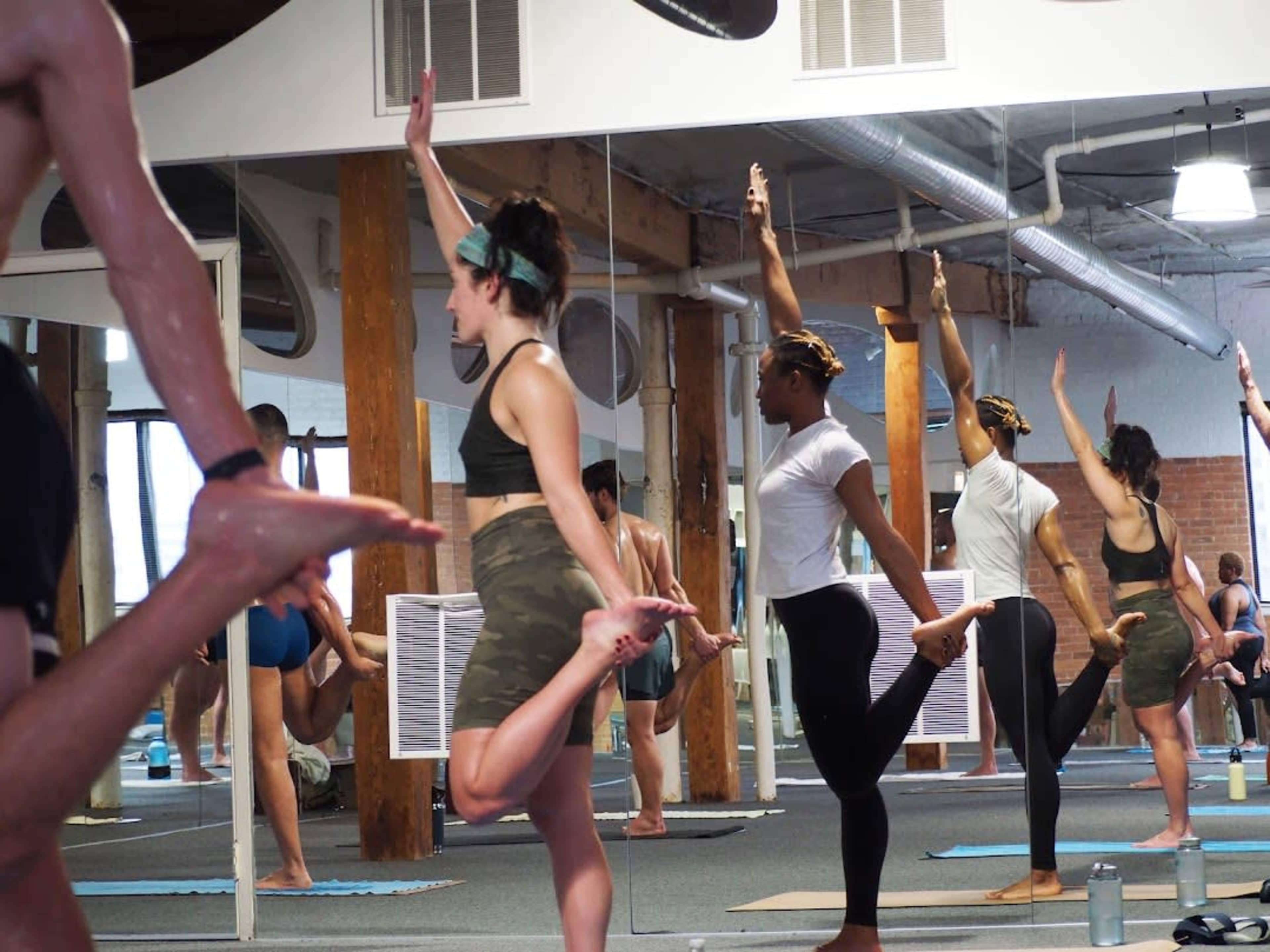 A group of individuals practices yoga in a studio with mirrors, balancing on one leg while holding their opposite foot.