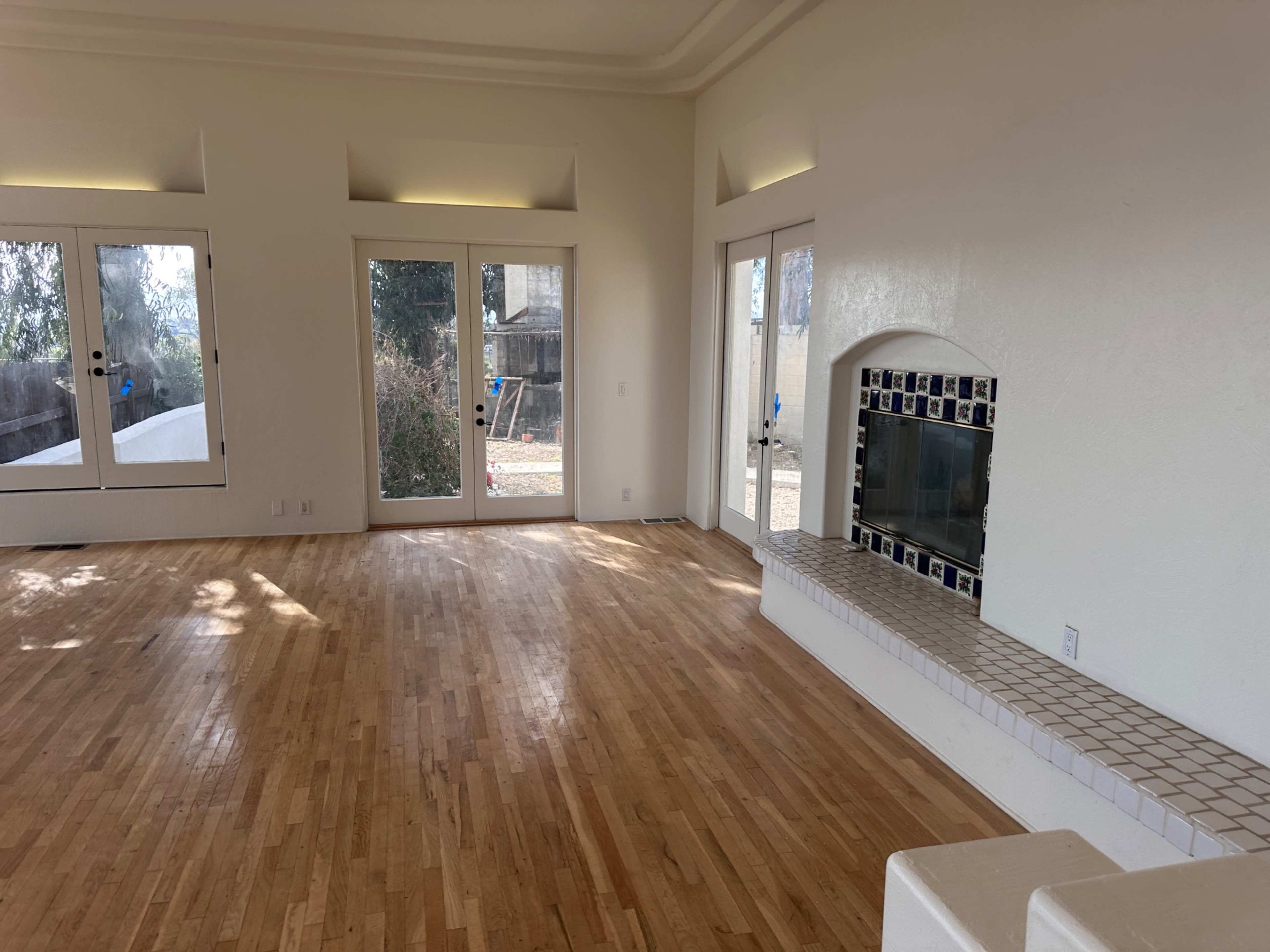 The image shows a spacious, empty living room with wooden flooring, a tiled fireplace, and large windows that open to a garden area.