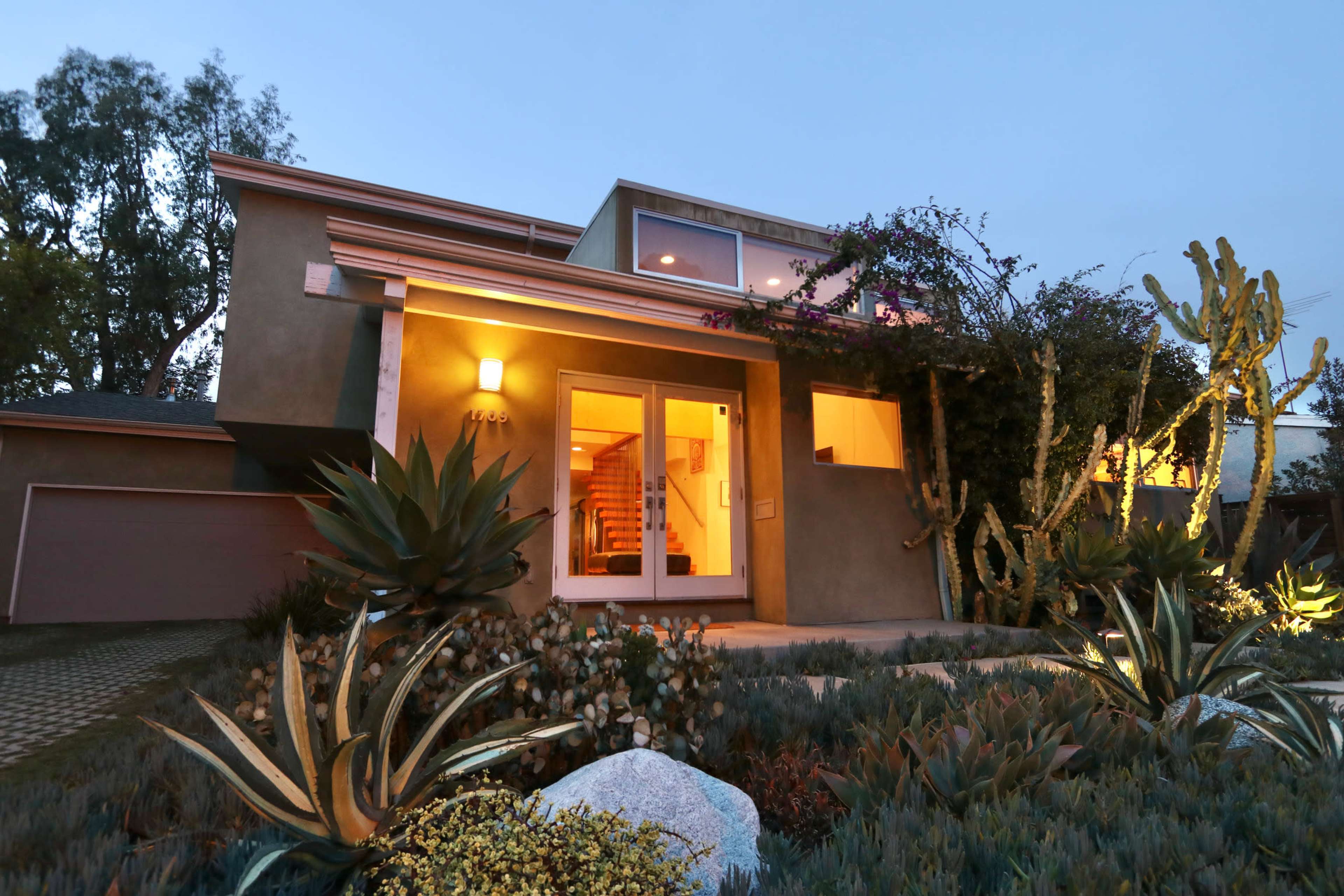 A modern house with large front windows, surrounded by various plants and illuminated by outdoor lighting in the evening.