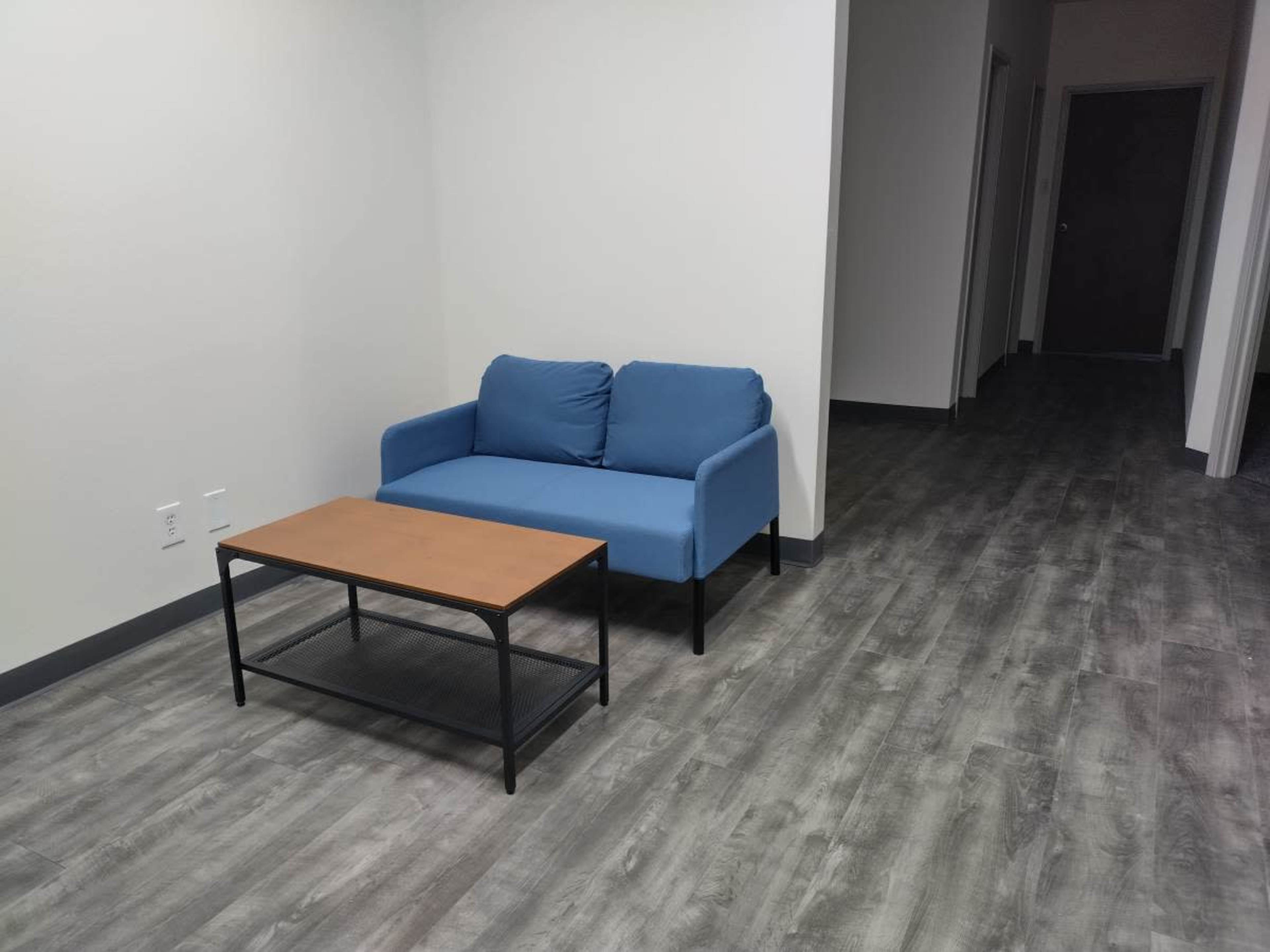 A blue sofa and a wooden coffee table are positioned in a sparse, well-lit room with a long hallway visible in the background.