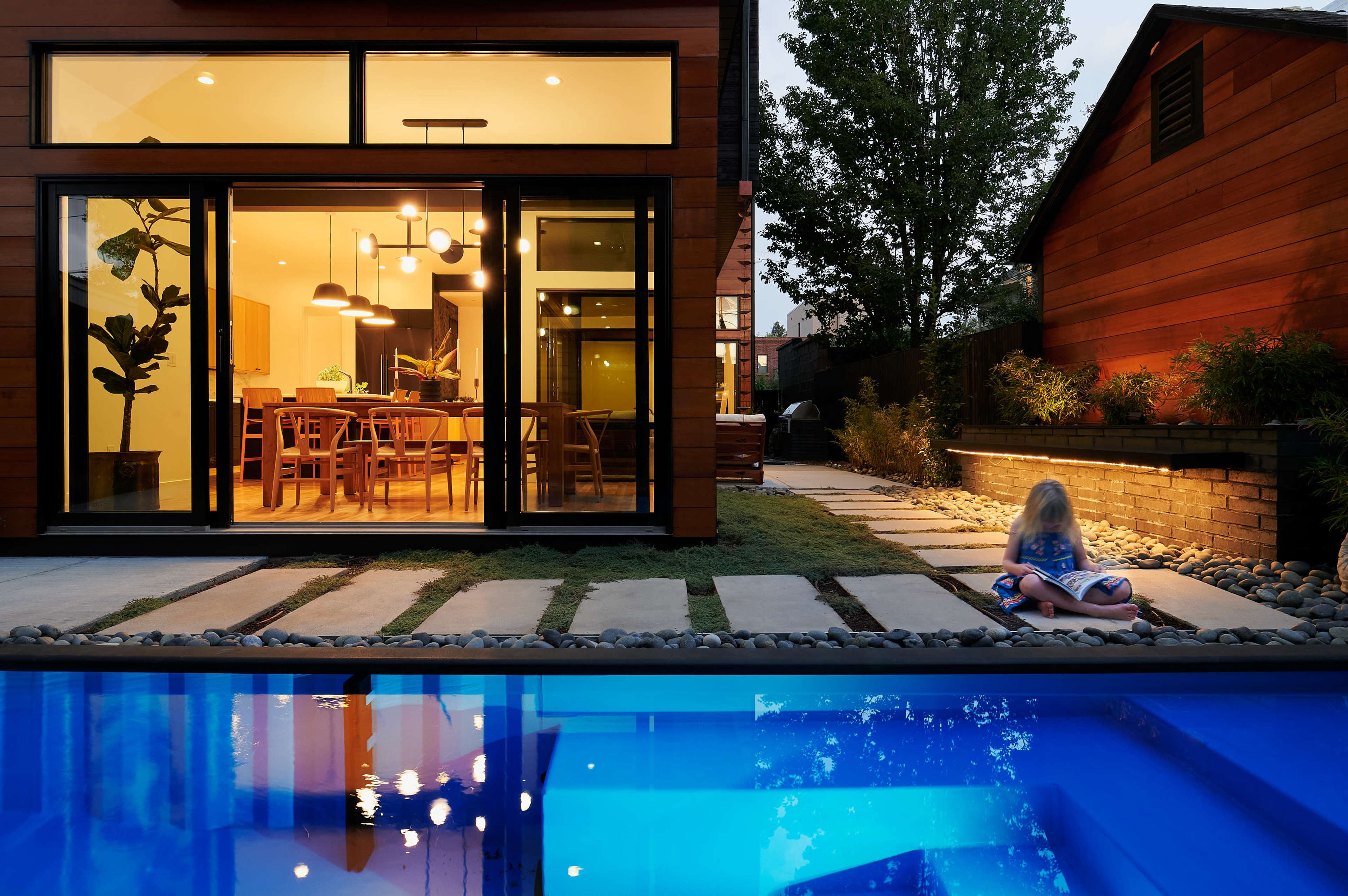 A child sits on a towel near a swimming pool in a backyard, with a modern house featuring large windows in the background.