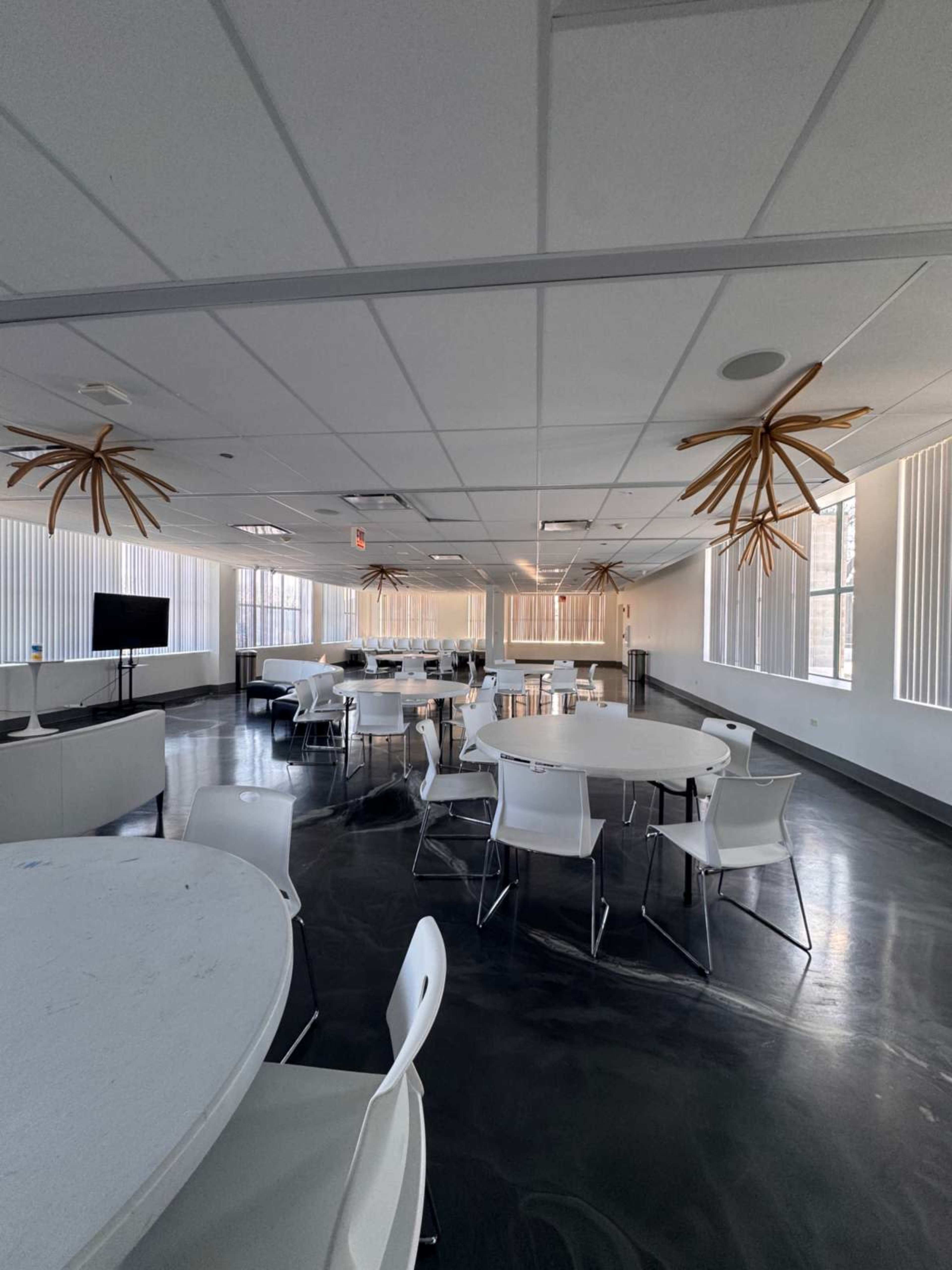 The image shows a spacious modern room with several round tables and chairs, along with unique light fixtures hanging from the ceiling.