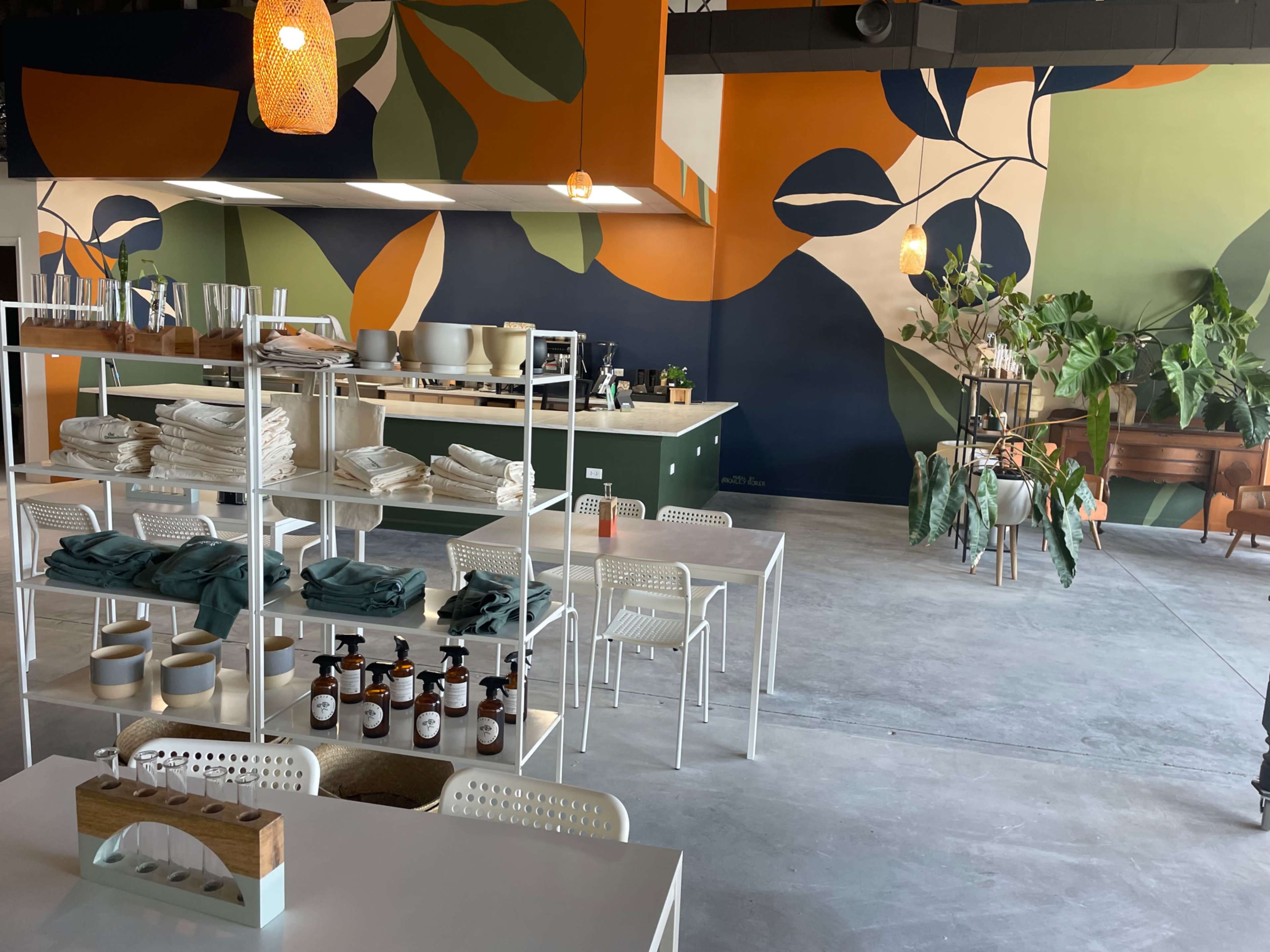The interior of a café features a colorful mural, white furniture, and shelves displaying neatly arranged items and plants.
